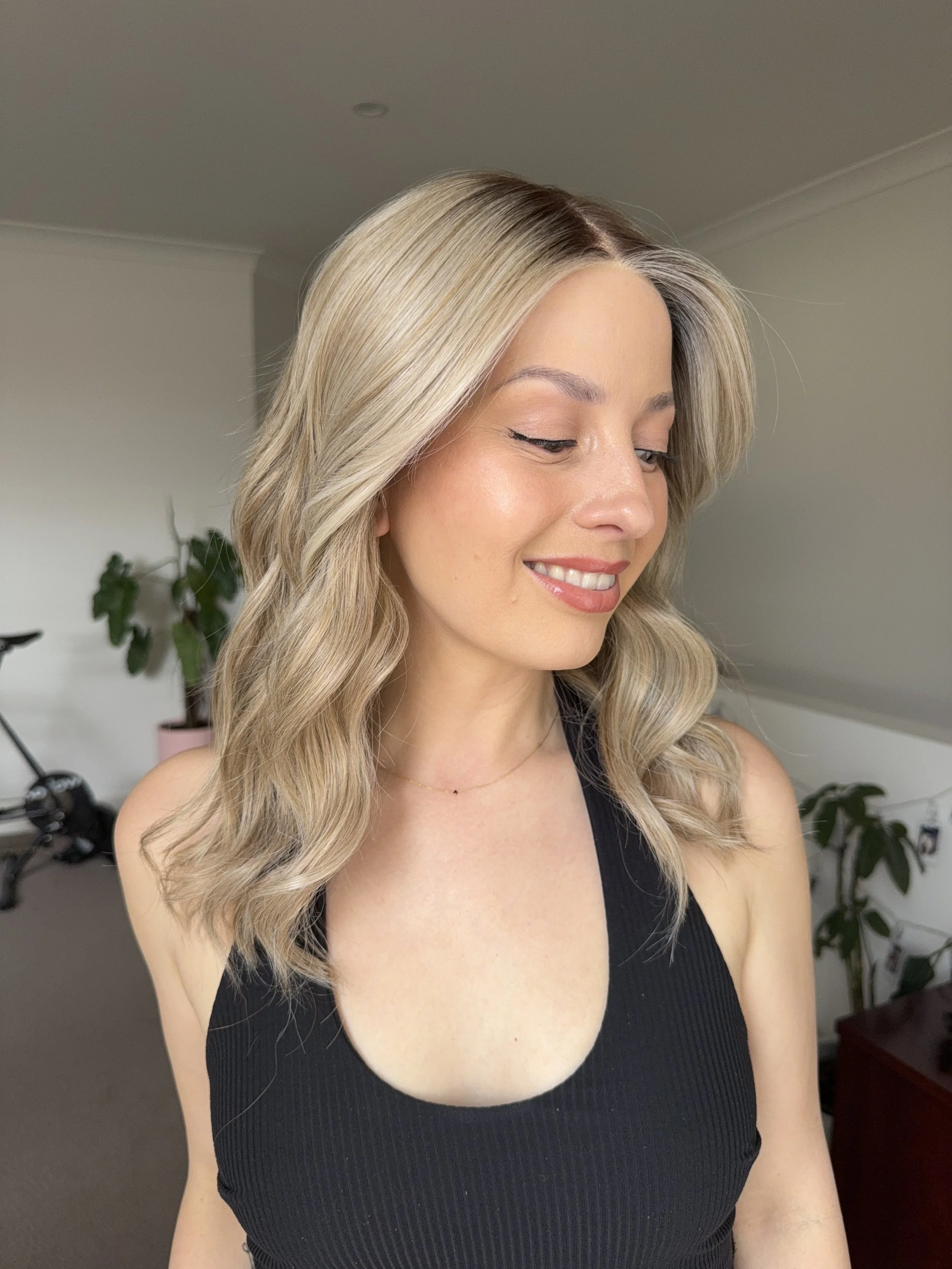 A woman with blonde, wavy hair wearing a black sleeveless top, smiling and looking down, in a room with a plant and a bicycle in the background.