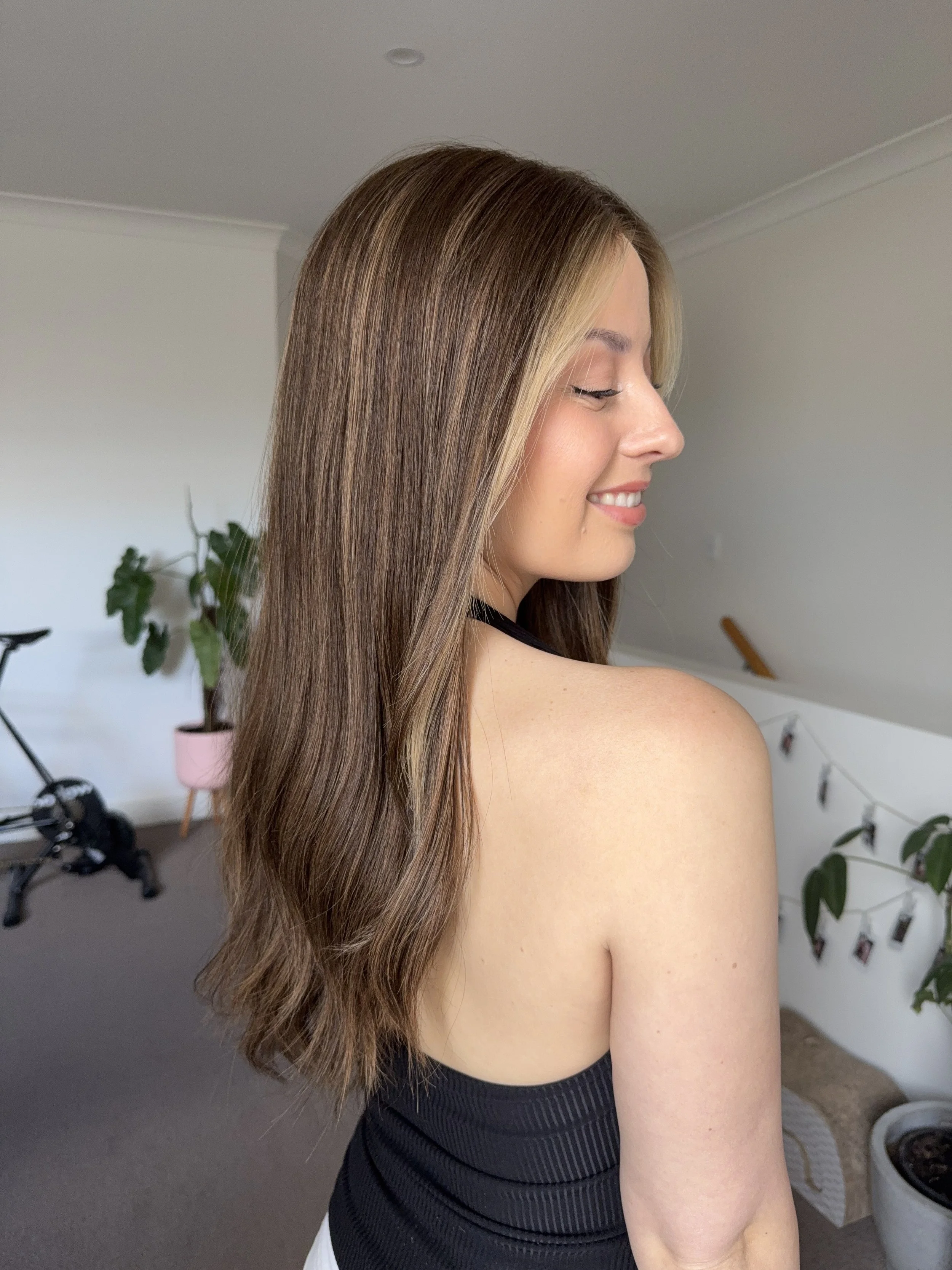 A woman with long, wavy brown hair and a light streak smiling and looking to the side in a bright room.