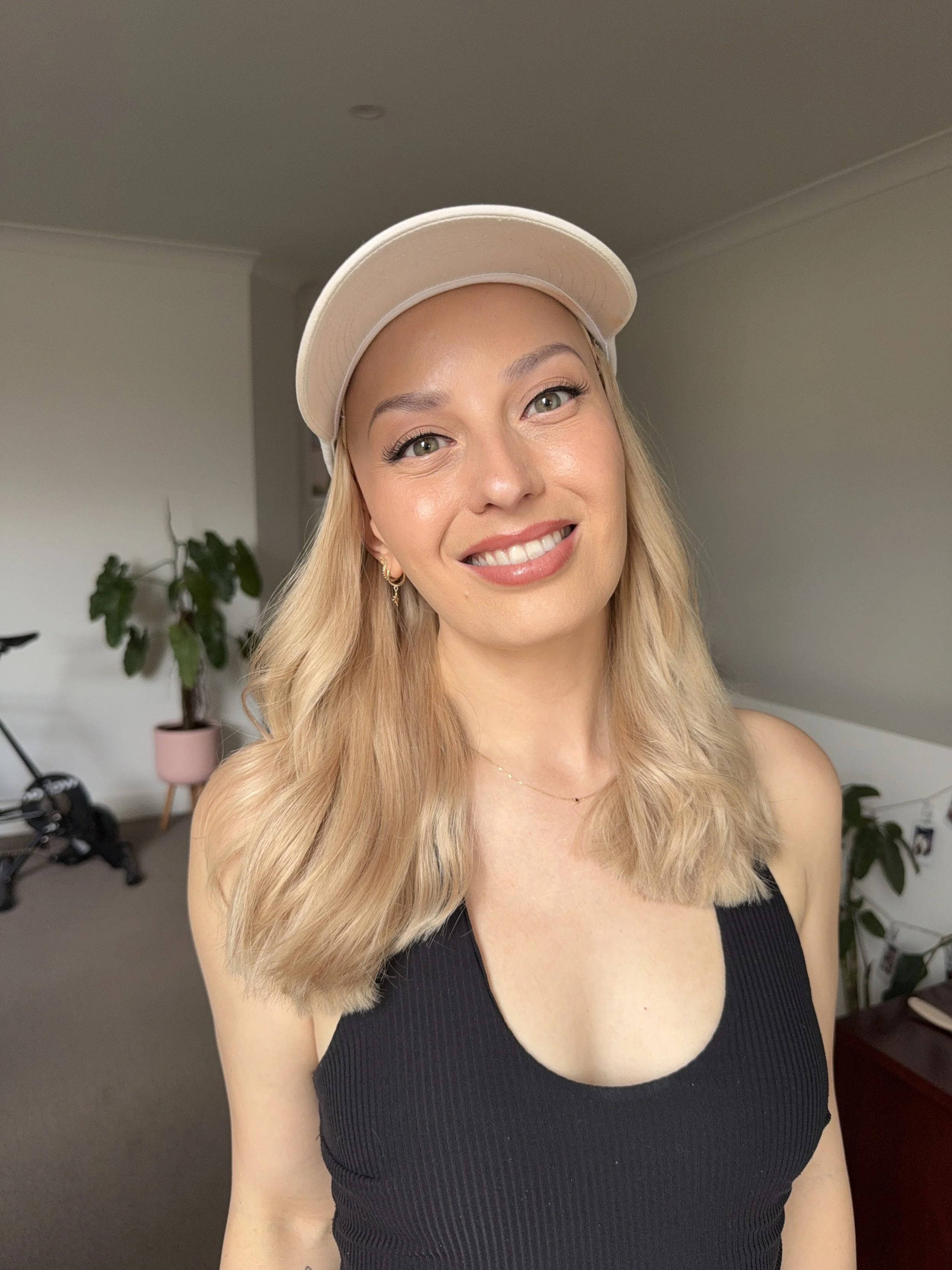 A young woman with blonde wavy hair wearing a beige baseball cap and a black sleeveless top, smiling indoors with a plant and bicycle in the background.
