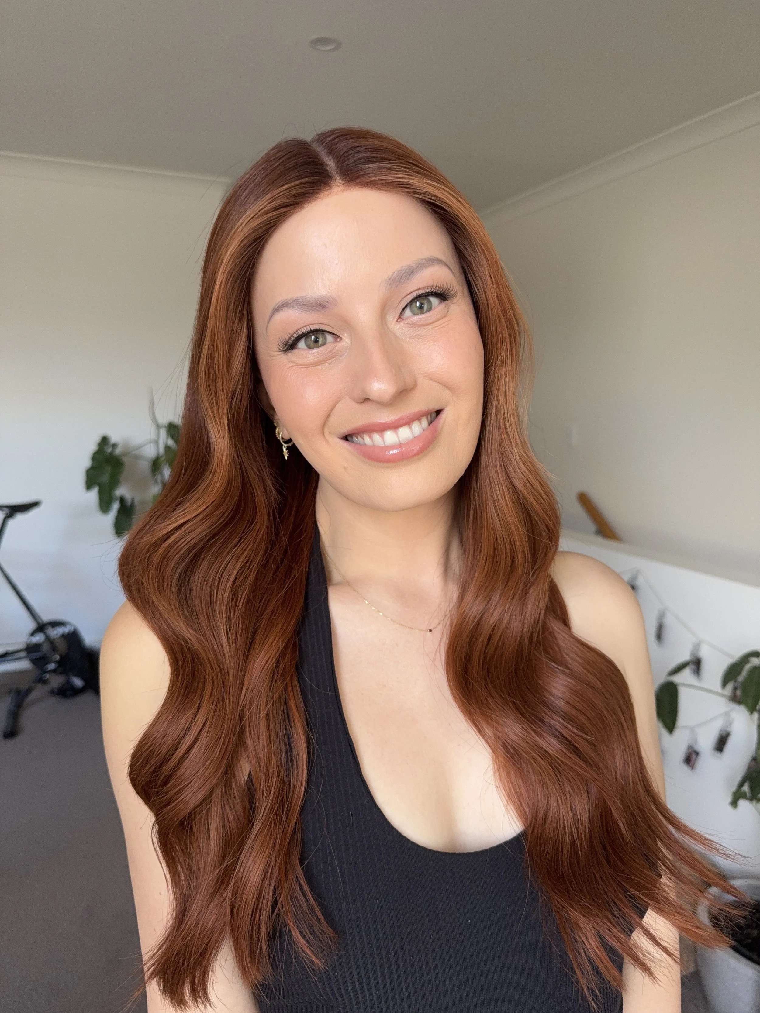 A smiling woman with long wavy red hair wearing a black sleeveless top, standing in a bright room with white walls and some green plants in the background.
