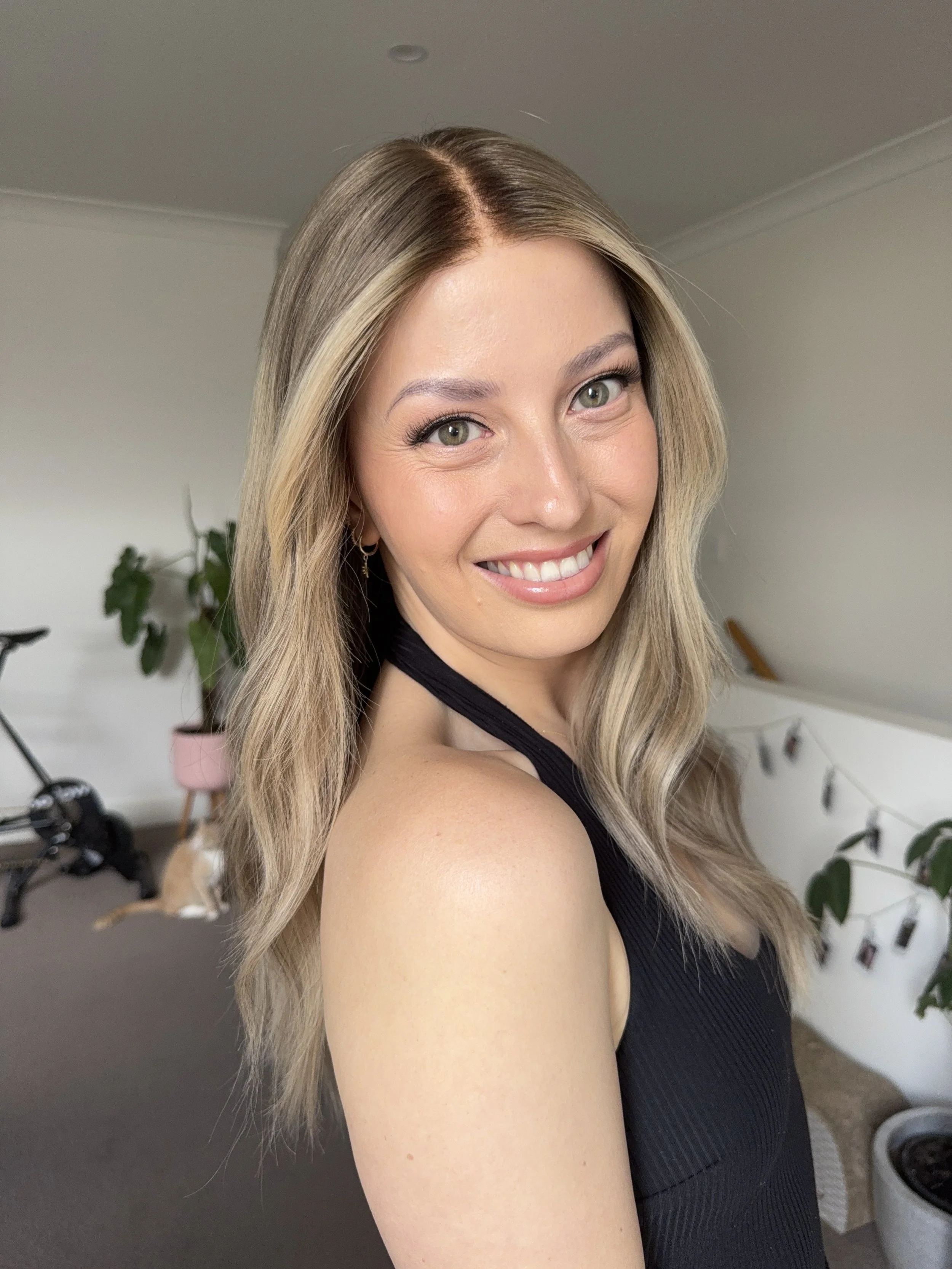 A woman with long wavy blonde hair, wearing a black sleeveless top, smiling, indoors with houseplants and a bicycle in the background.