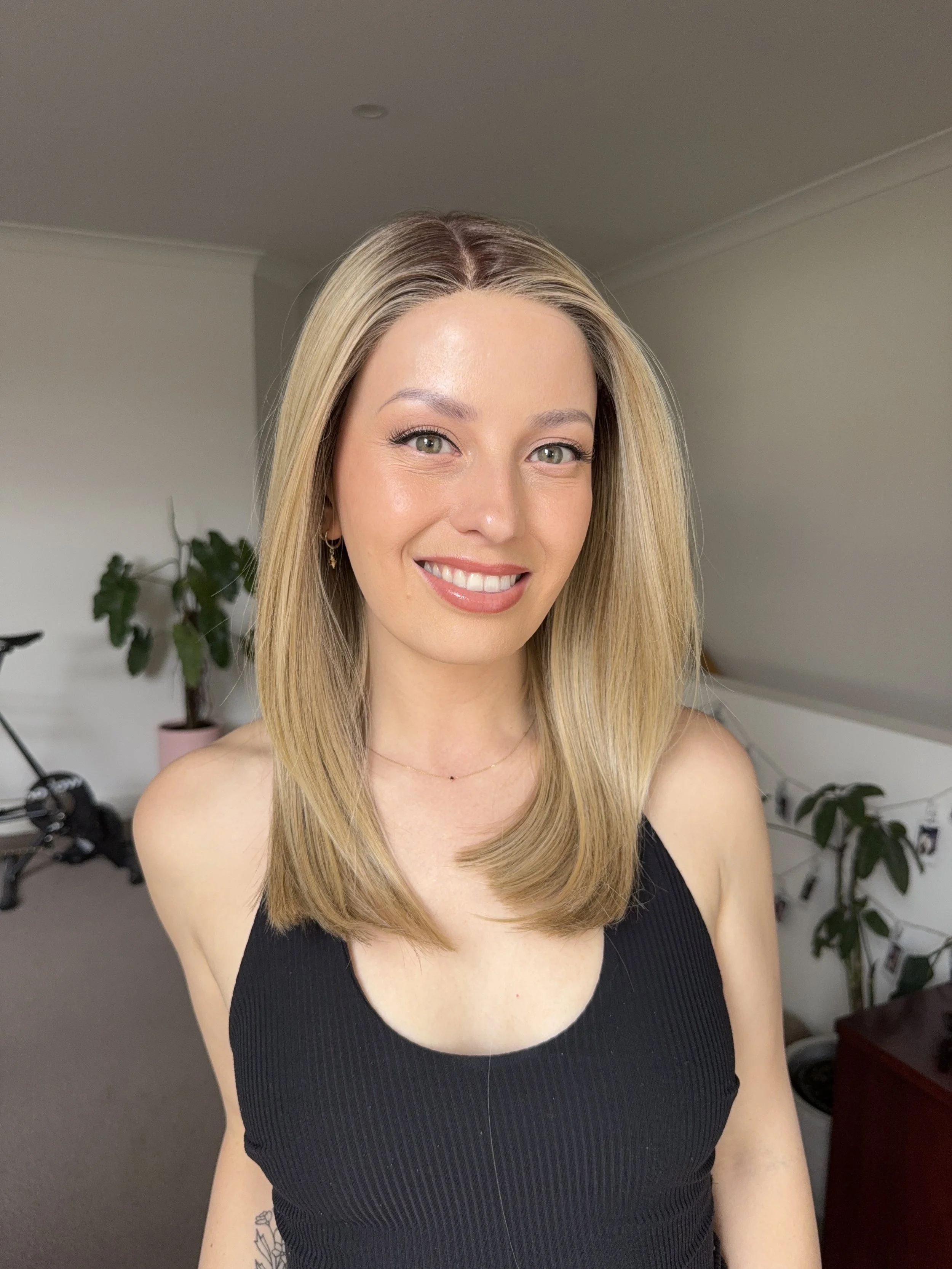 A smiling woman with blonde hair wearing a black tank top in a room with a plant, a bicycle, and string lights in the background.