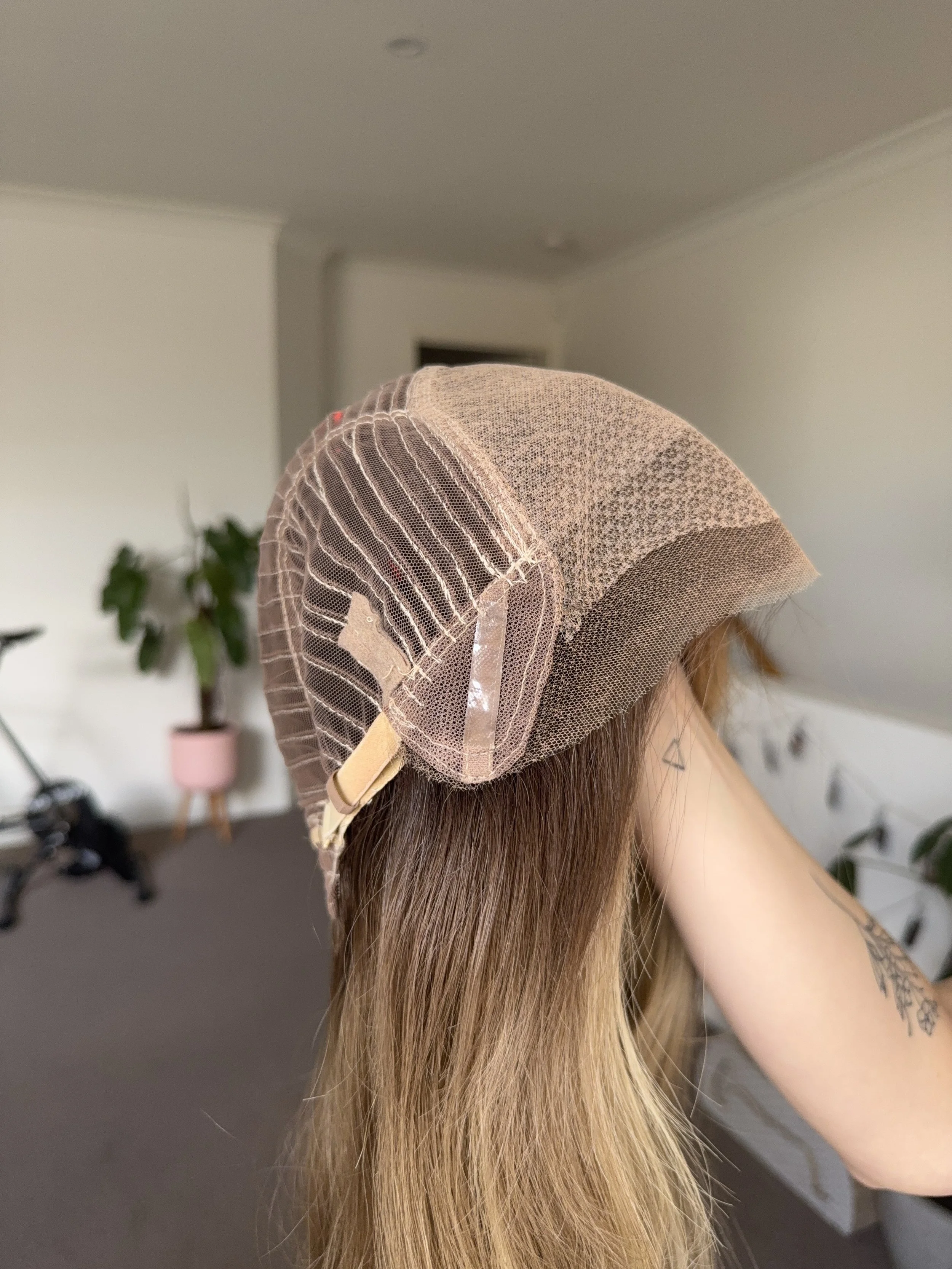 Patchwork wig cap on long, blond hair, inside a room with white walls and a potted plant in the background.