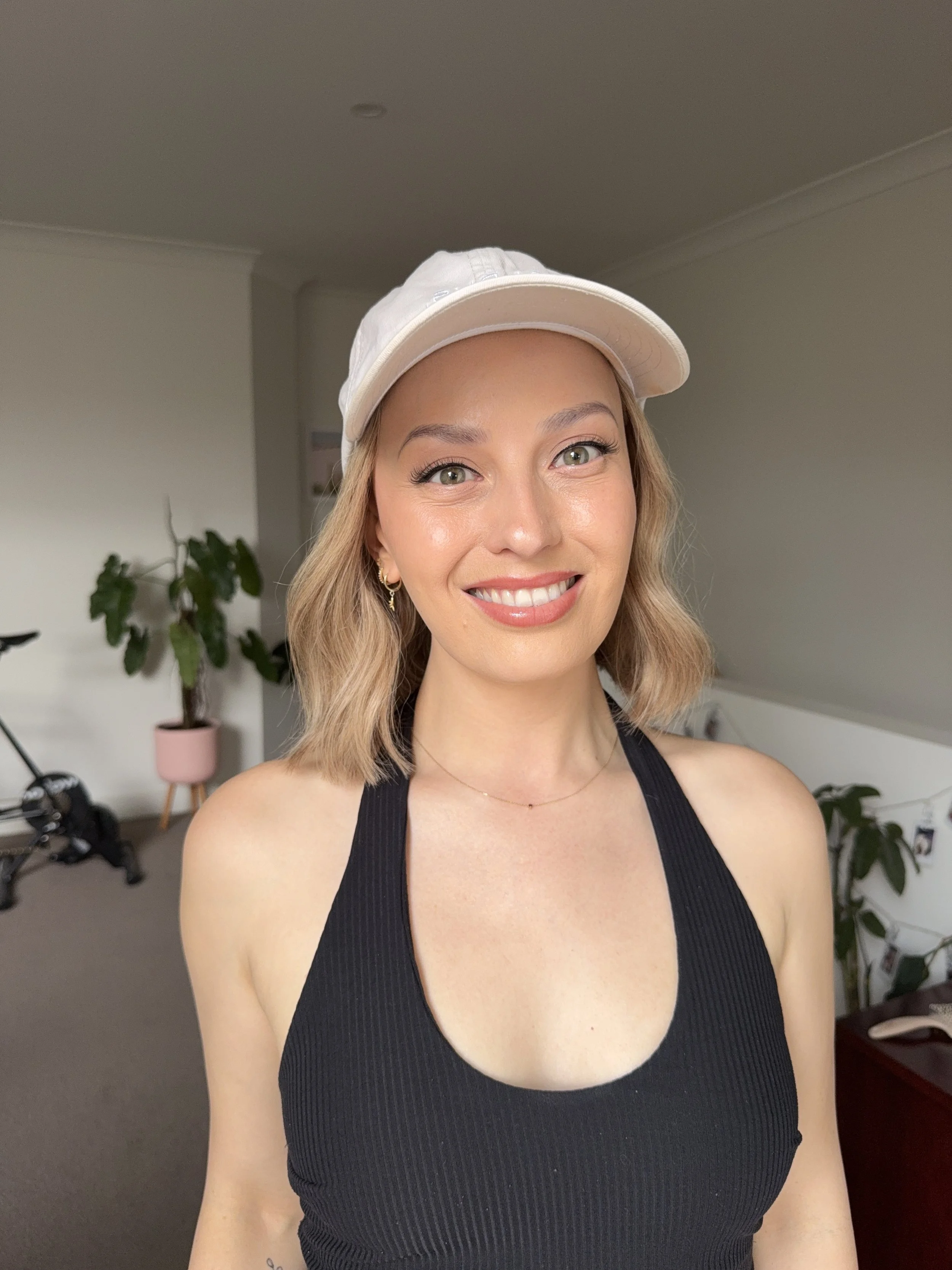 A young woman with blonde hair, wearing a beige cap and a black tank top, smiling indoors with a houseplant and bicycle in the background.