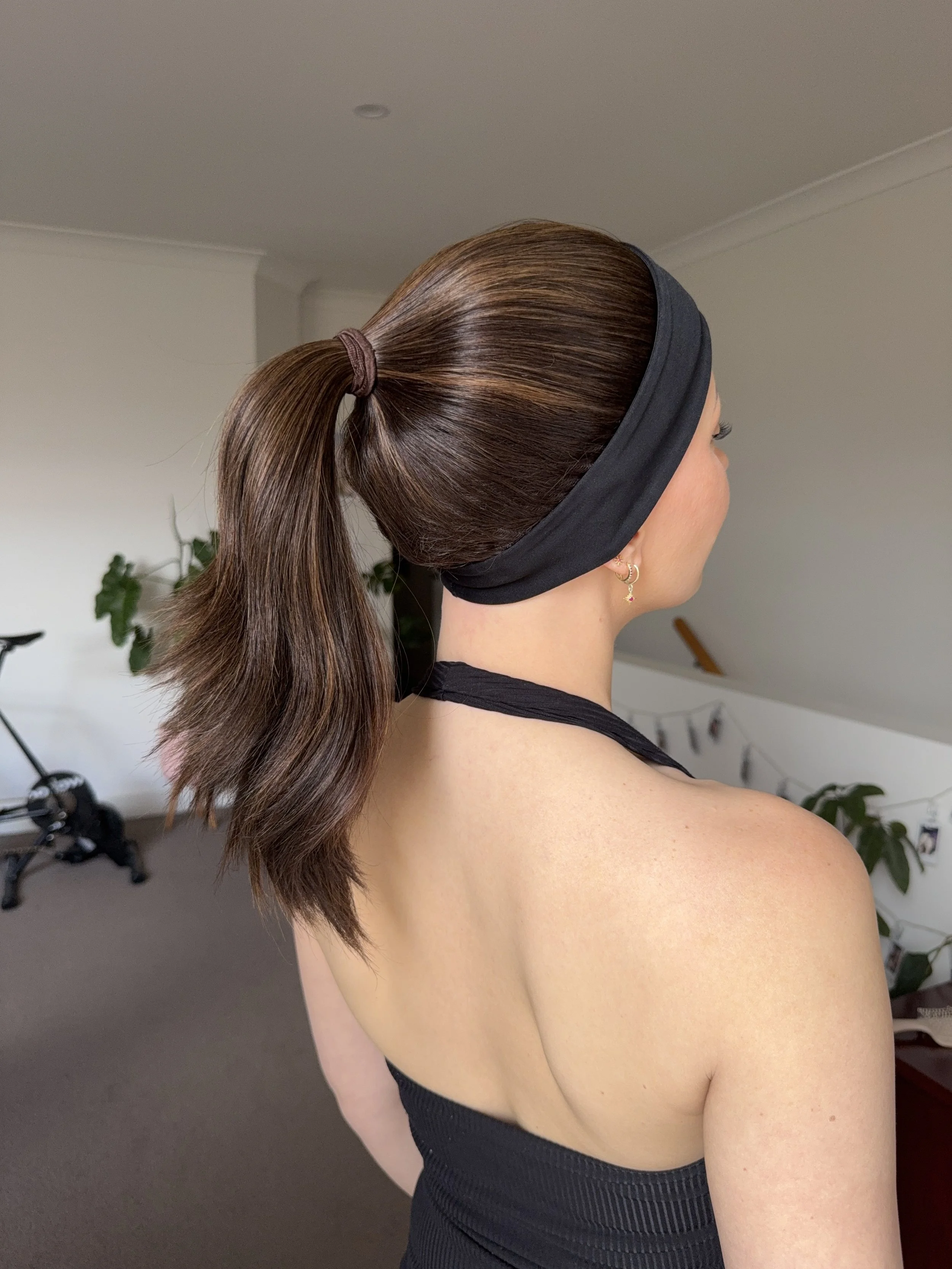 A woman with long brown hair tied in a ponytail, wearing a black headband, facing sideways in a room with a bicycle and plants in the background.