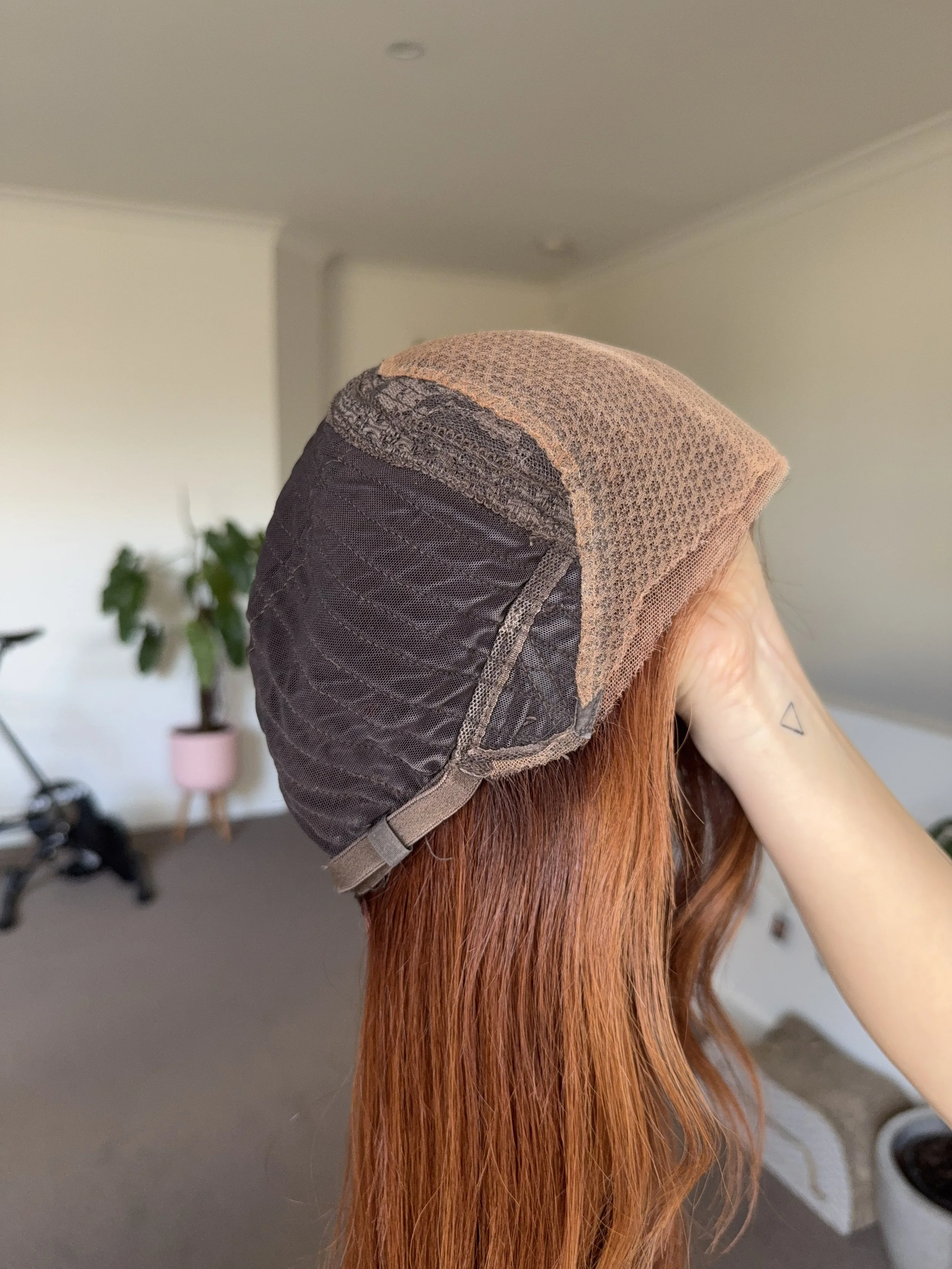 Close-up view of a person with long red hair wearing a wig cap, held by a hand with a small triangle tattoo on the wrist, indoors with a plant and furniture in the background.