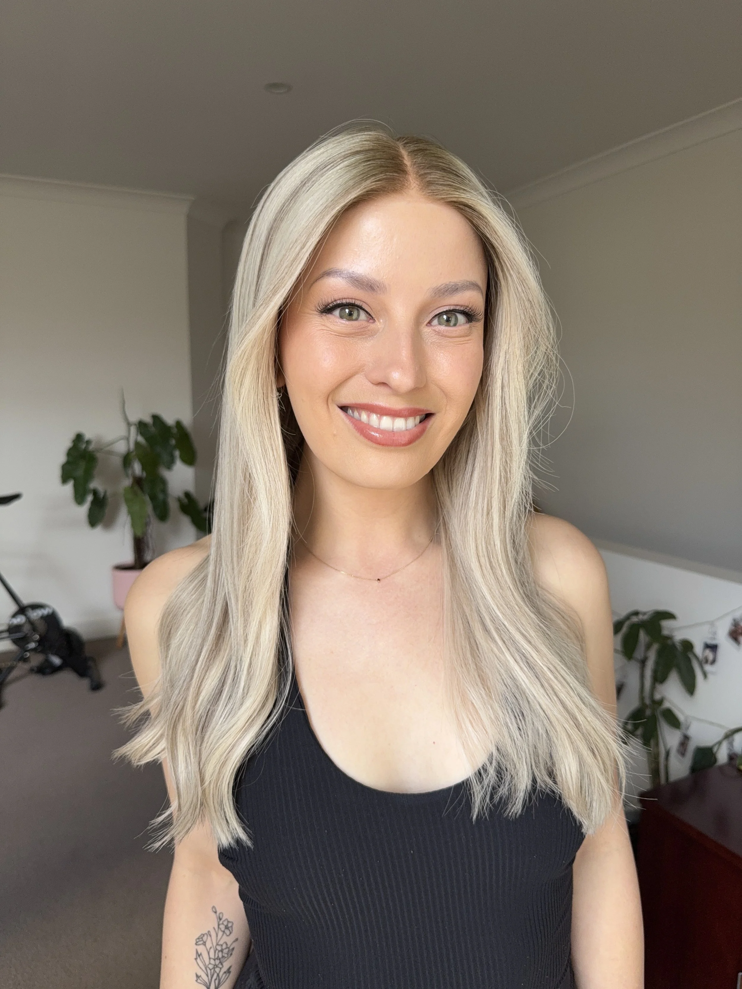 A woman with long blonde hair, green eyes, and a bright smile wearing a black sleeveless top, standing in a room with houseplants and a neutral-colored background.