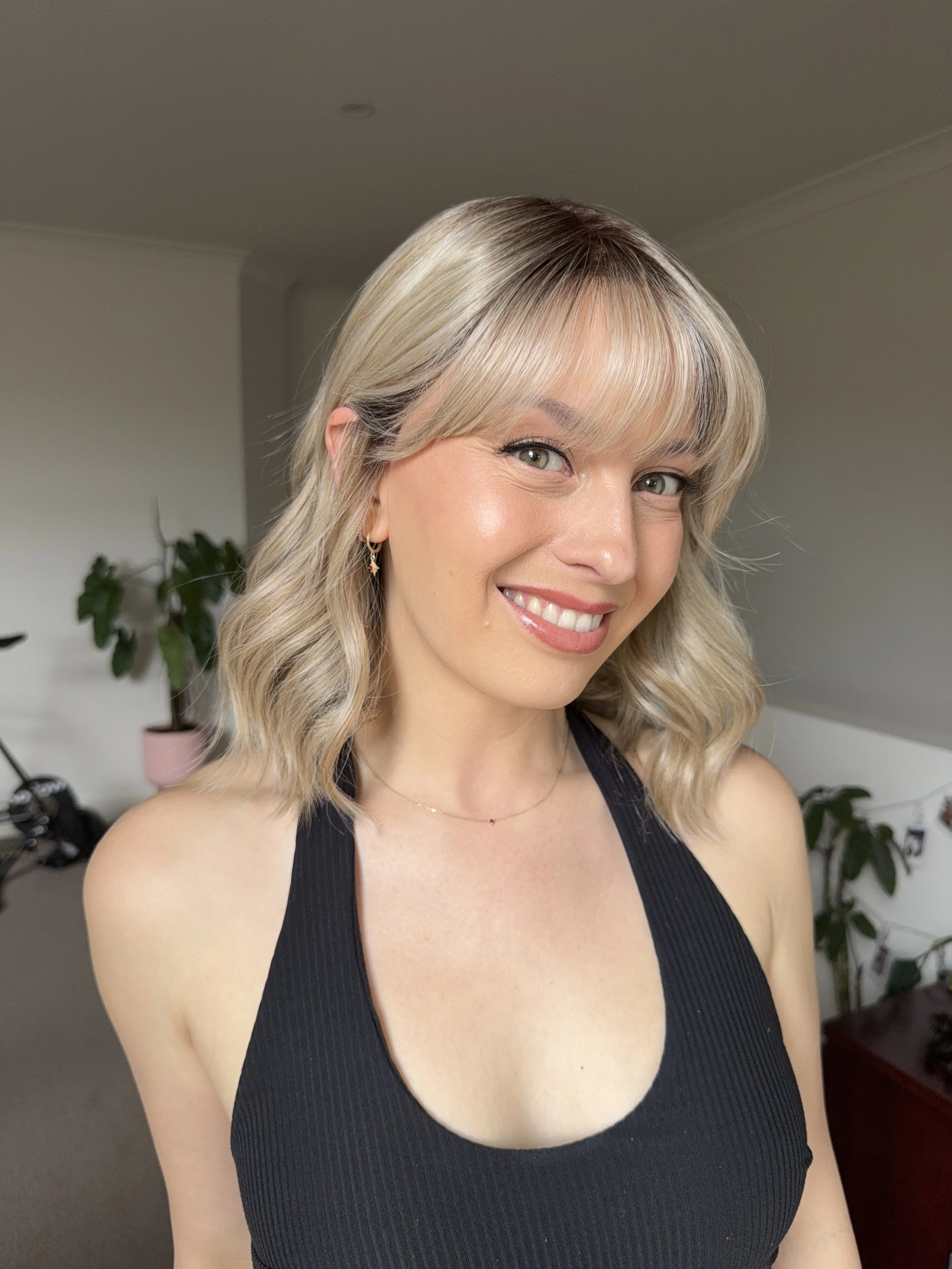 A woman with blonde wavy hair smiling in a room with houseplants and furniture.