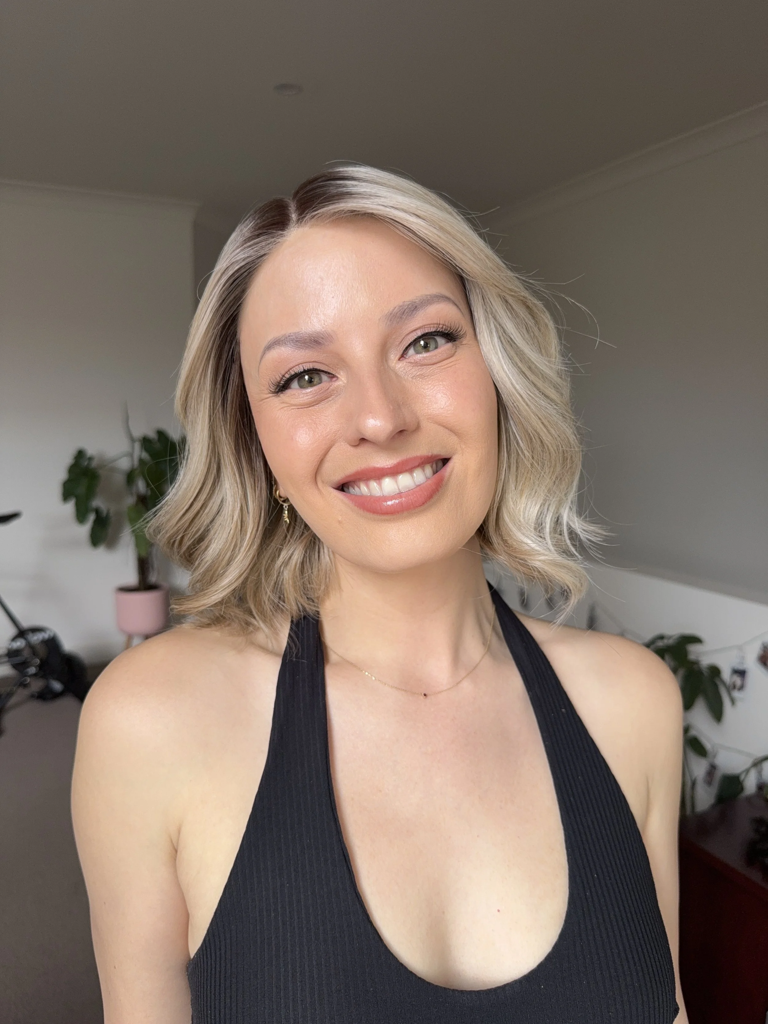 A smiling woman with wavy blonde hair wearing a black tank top, standing indoors with plants and furniture in the background.