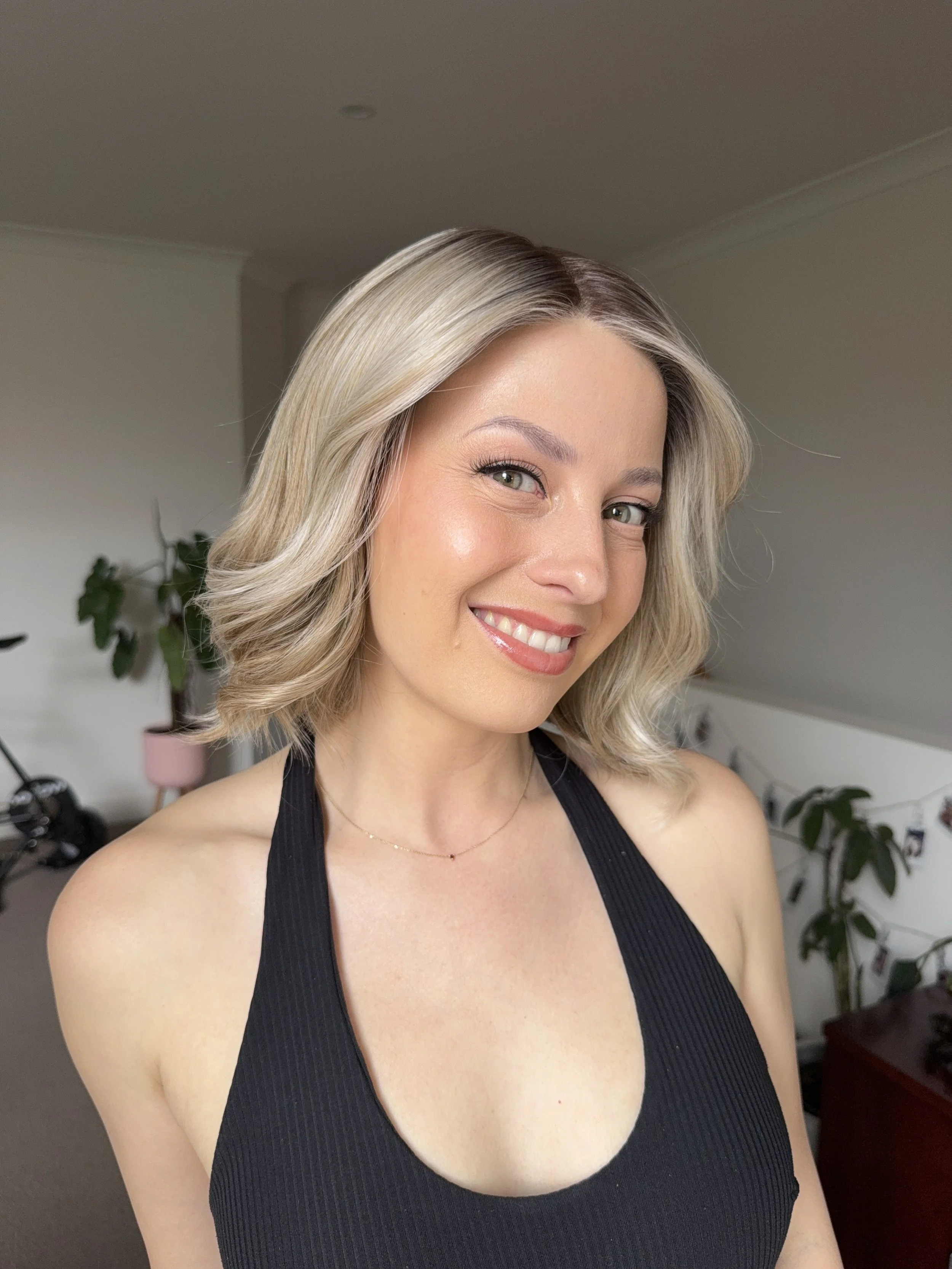 Close-up of a smiling woman with shoulder-length blonde hair, wearing a black tank top, indoors with a plant and furniture in the background.
