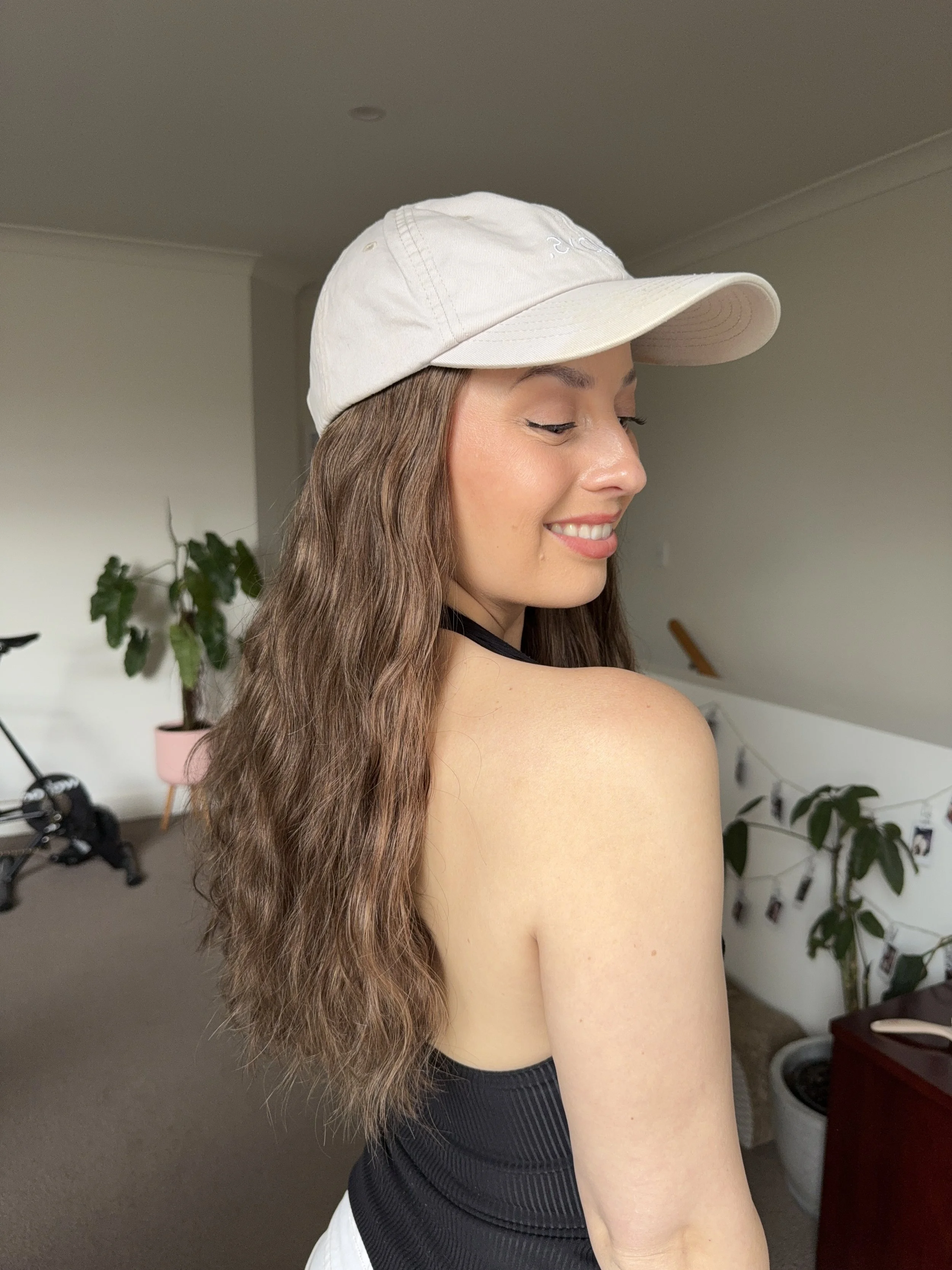 A young woman with long wavy brown hair, wearing a beige baseball cap, black tank top, and light-colored bottom, smiling with her eyes closed in a cozy indoor setting with plants and photos.