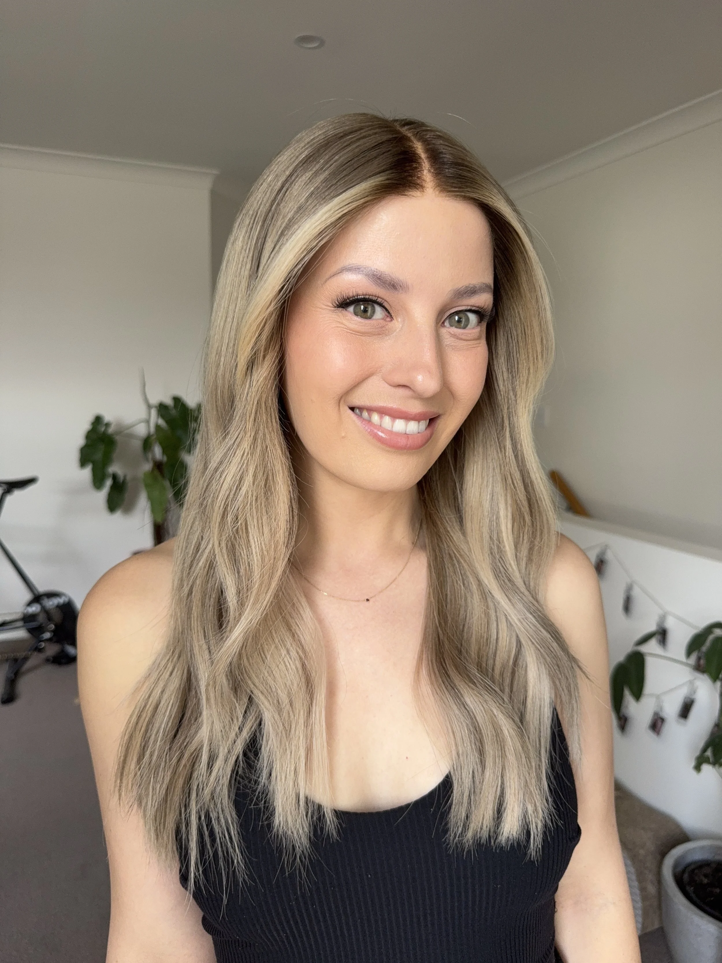 A young woman with long, wavy blond hair and green eyes smiling at the camera, wearing a black sleeveless top in a room with some plants and furniture in the background.