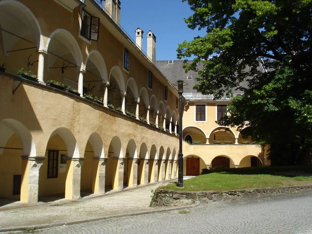 Ein gelbes Gebäude mit mehreren Bögen und einer Terrasse, umgeben von Bäumen und einem Gehweg, bei sonnigem Wetter.