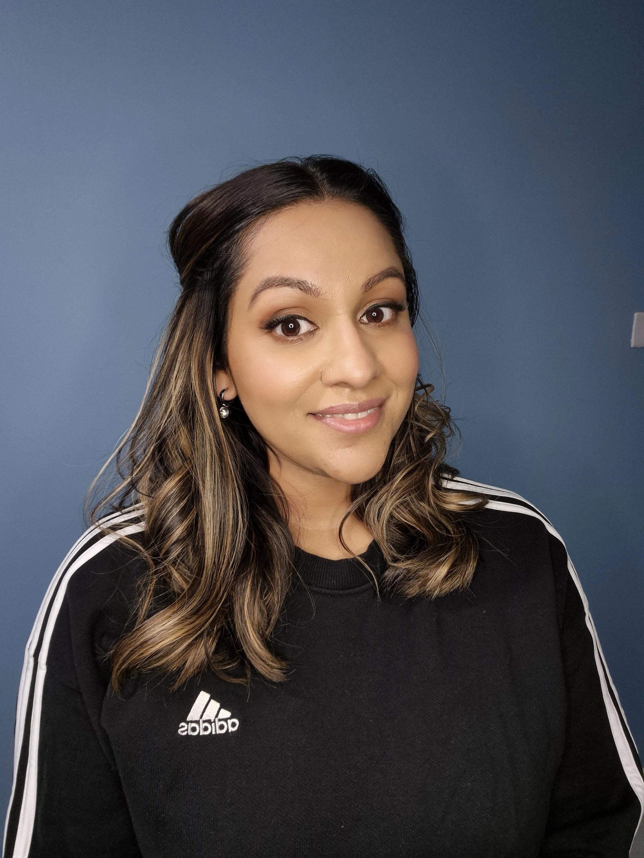 A woman with shoulder-length wavy hair, wearing a black Borussia Dortmund sweatshirt with white stripes on the sleeves, standing against a blue background.
