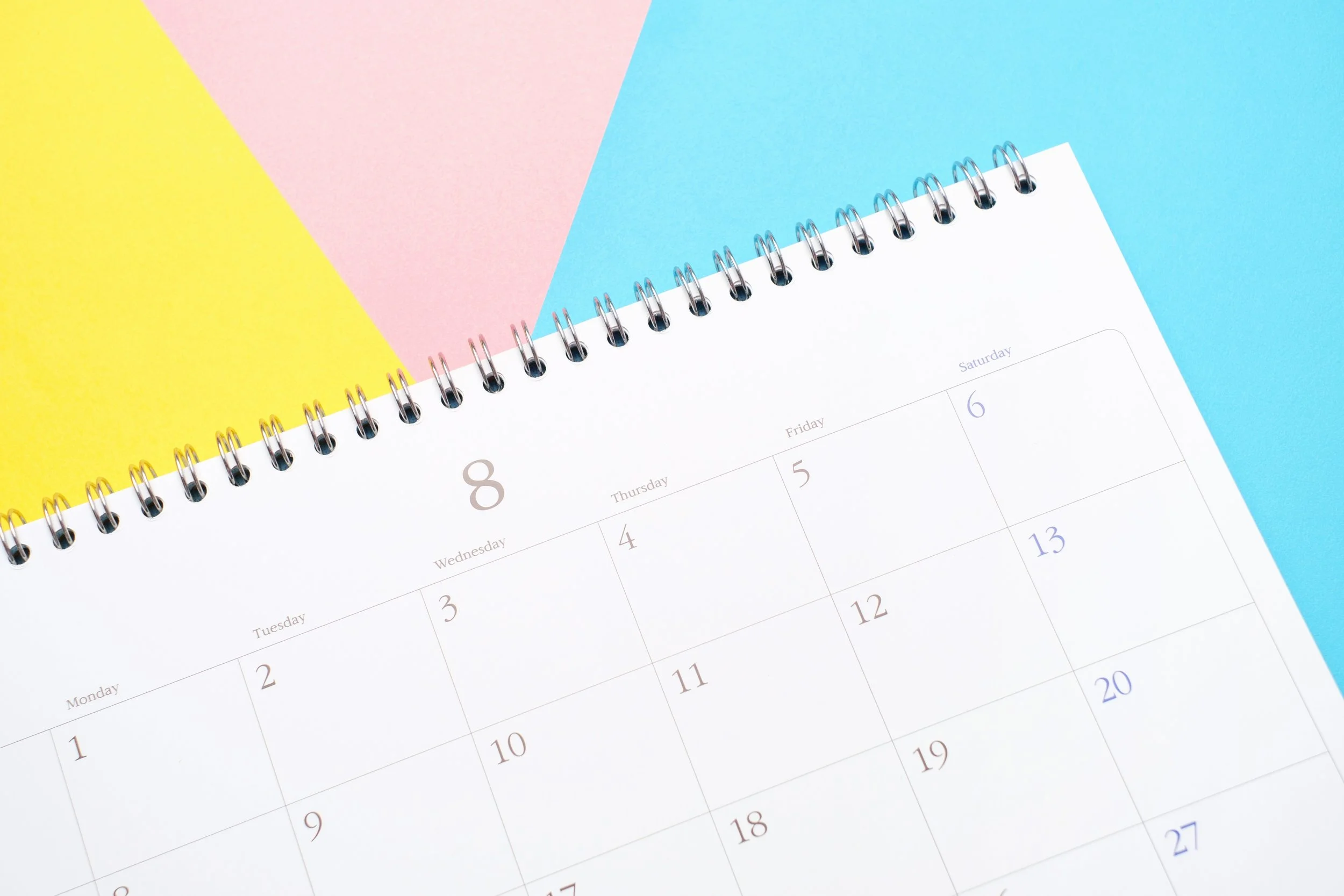 A spiral-bound calendar open to August, with visible dates and days, placed on a colorful background of yellow, pink, and blue paper.