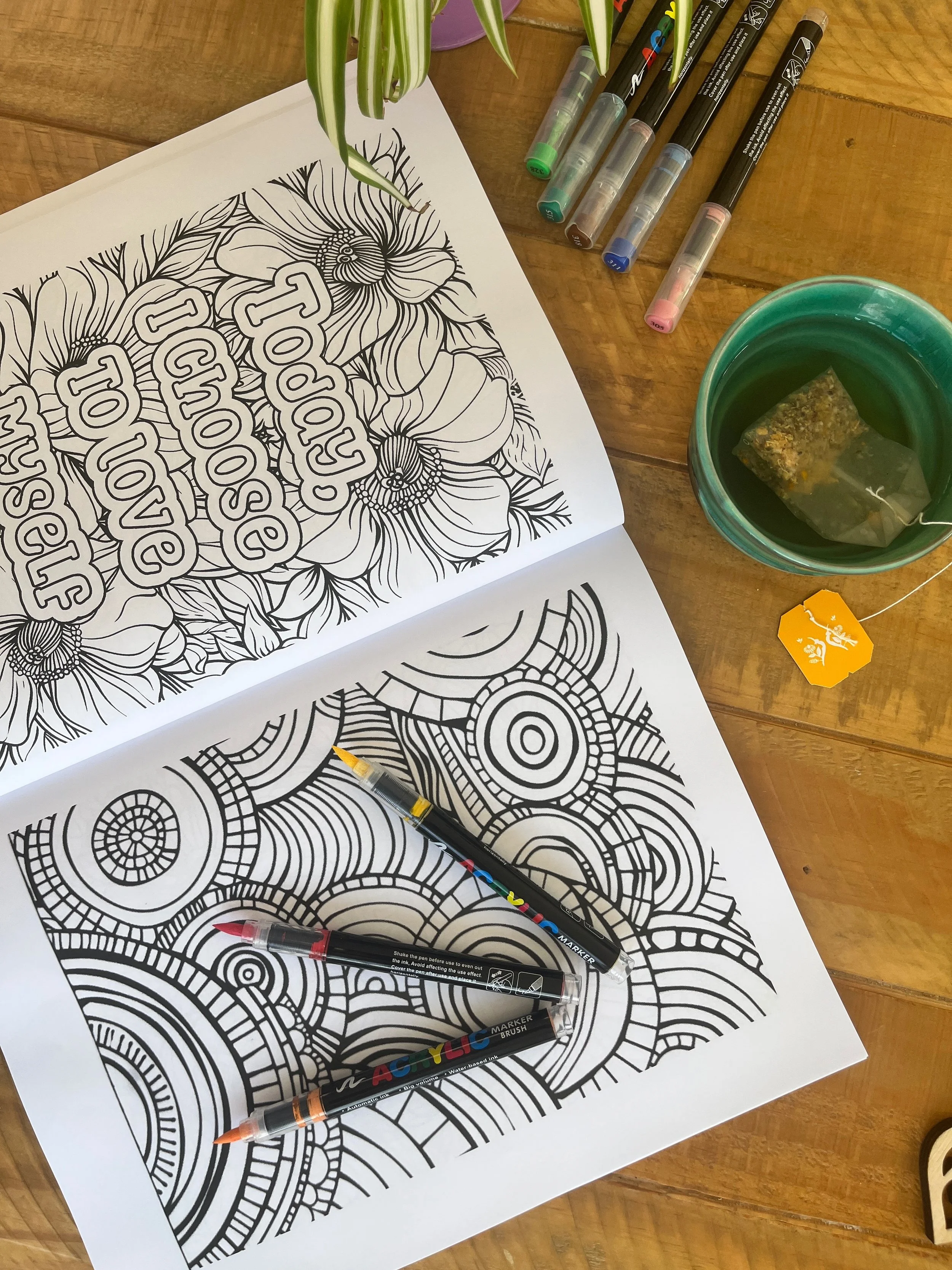 Coloring books with intricate black and white patterns, markers, a cup of tea with a tea bag, and a yellow tea tag on a wooden table.