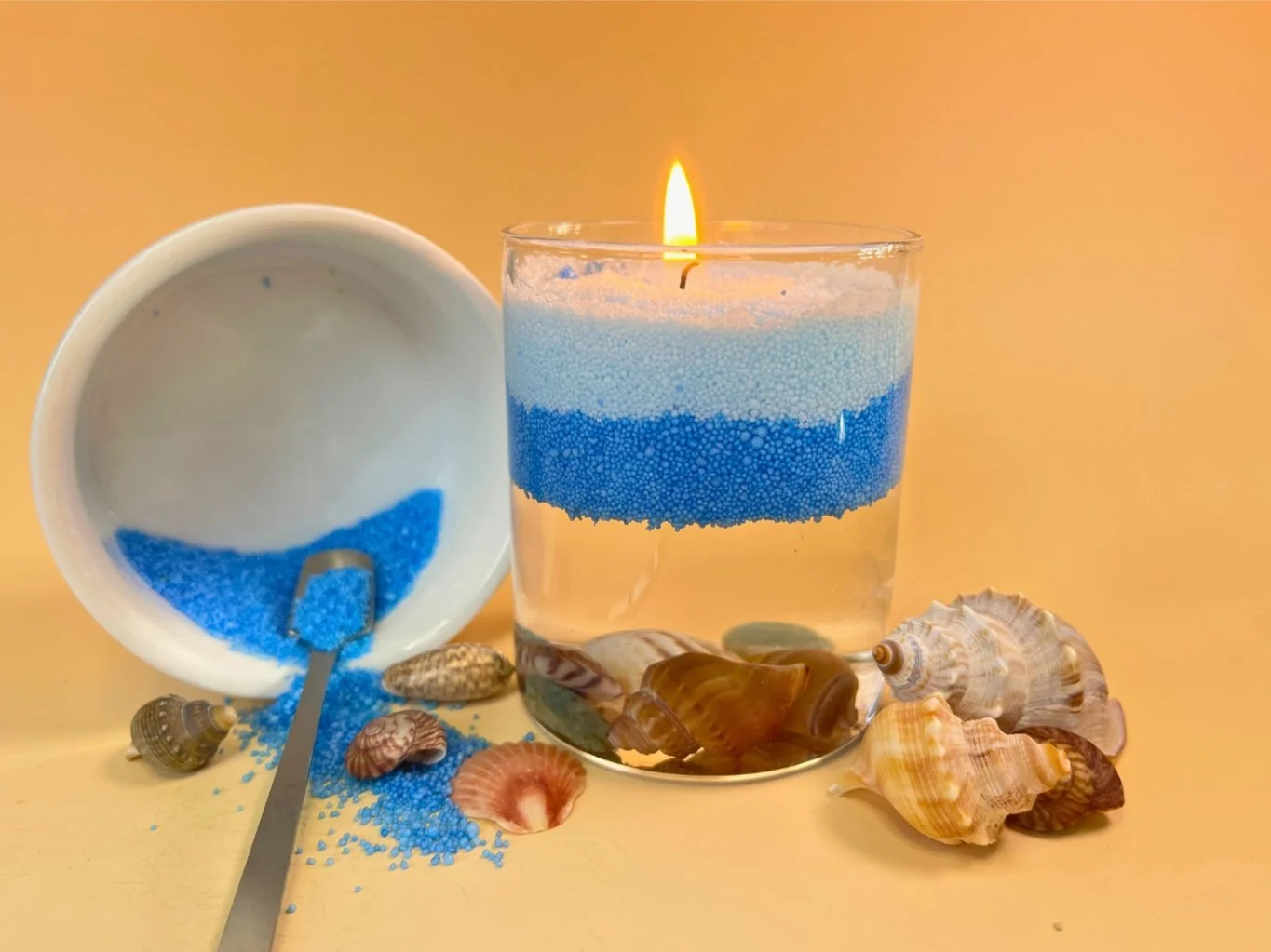 A lit candle with blue and white layers of wax in a glass container, seashells surrounding it, and an open container with blue bath salts spilling out, on a yellow surface.