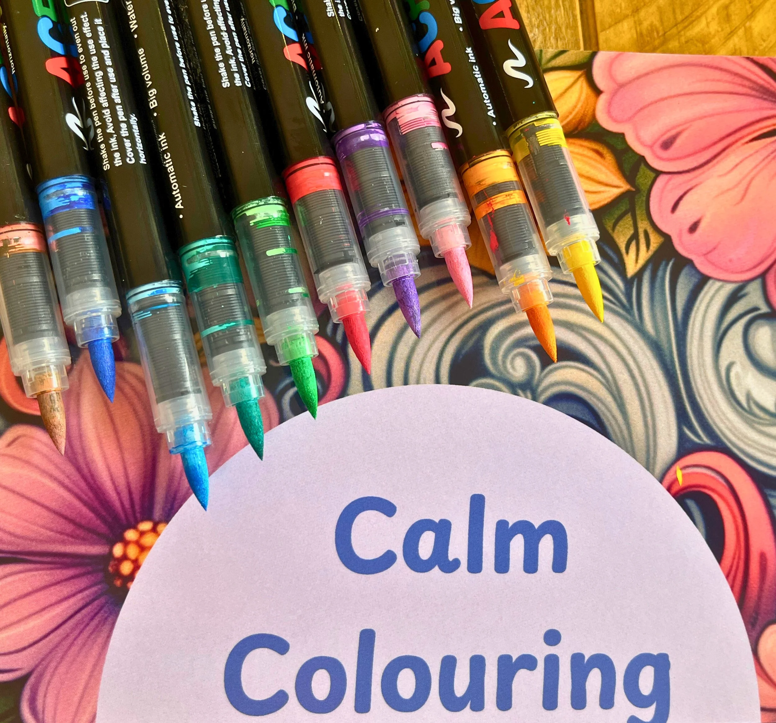 A variety of colorful markers with fine tips arranged on a floral patterned surface, partially covering a round purple paper with the words "Calm Colouring" written on it.