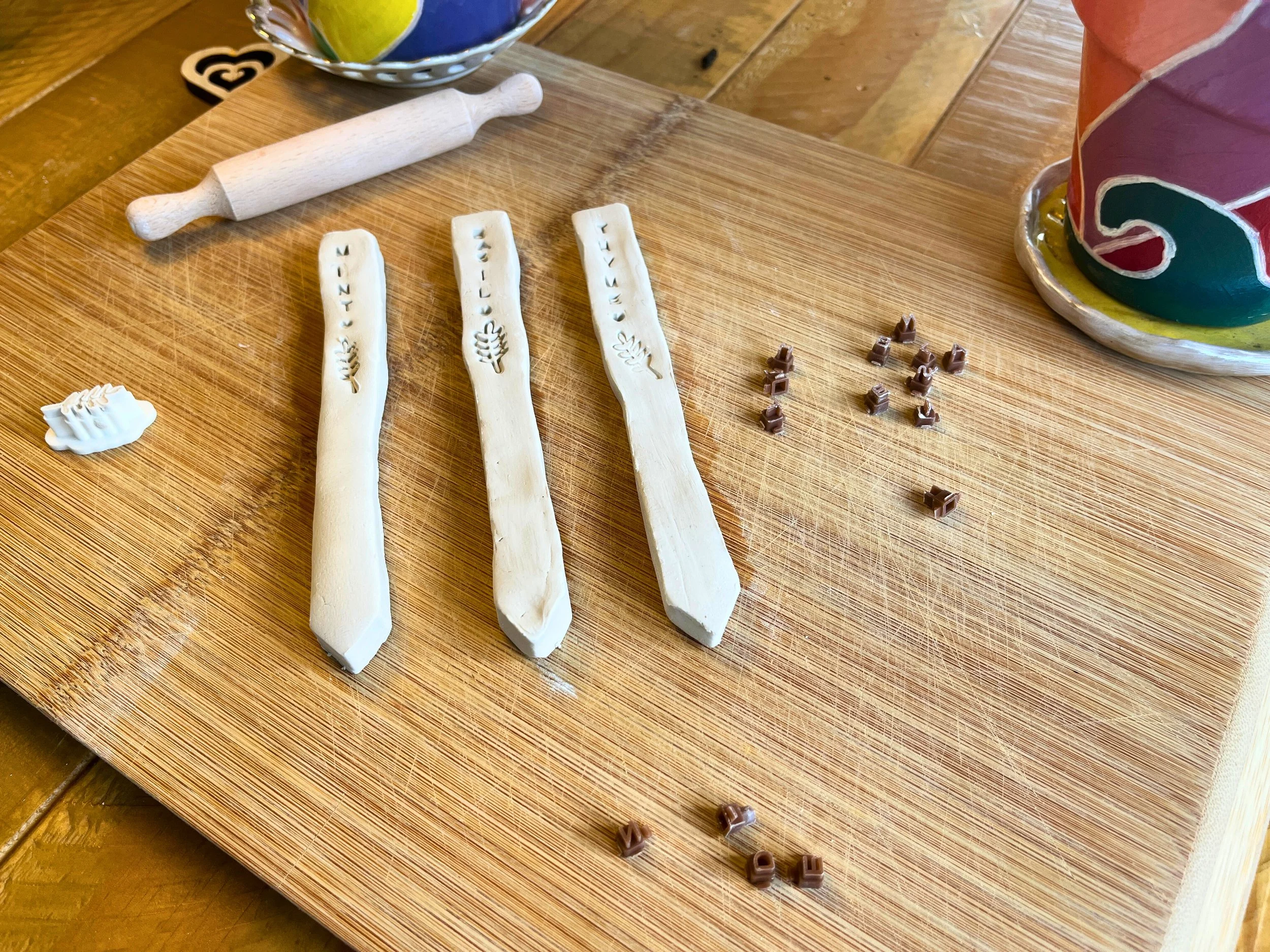Four carved wooden sticks with the words 'ZIT', 'HELLO', 'FELINE', and 'YELLOW' decorated with small engraved designs, a small white molded decorative piece, a wooden rolling pin, a colorful ceramic pot, and small brown alphabet beads on a wooden cut