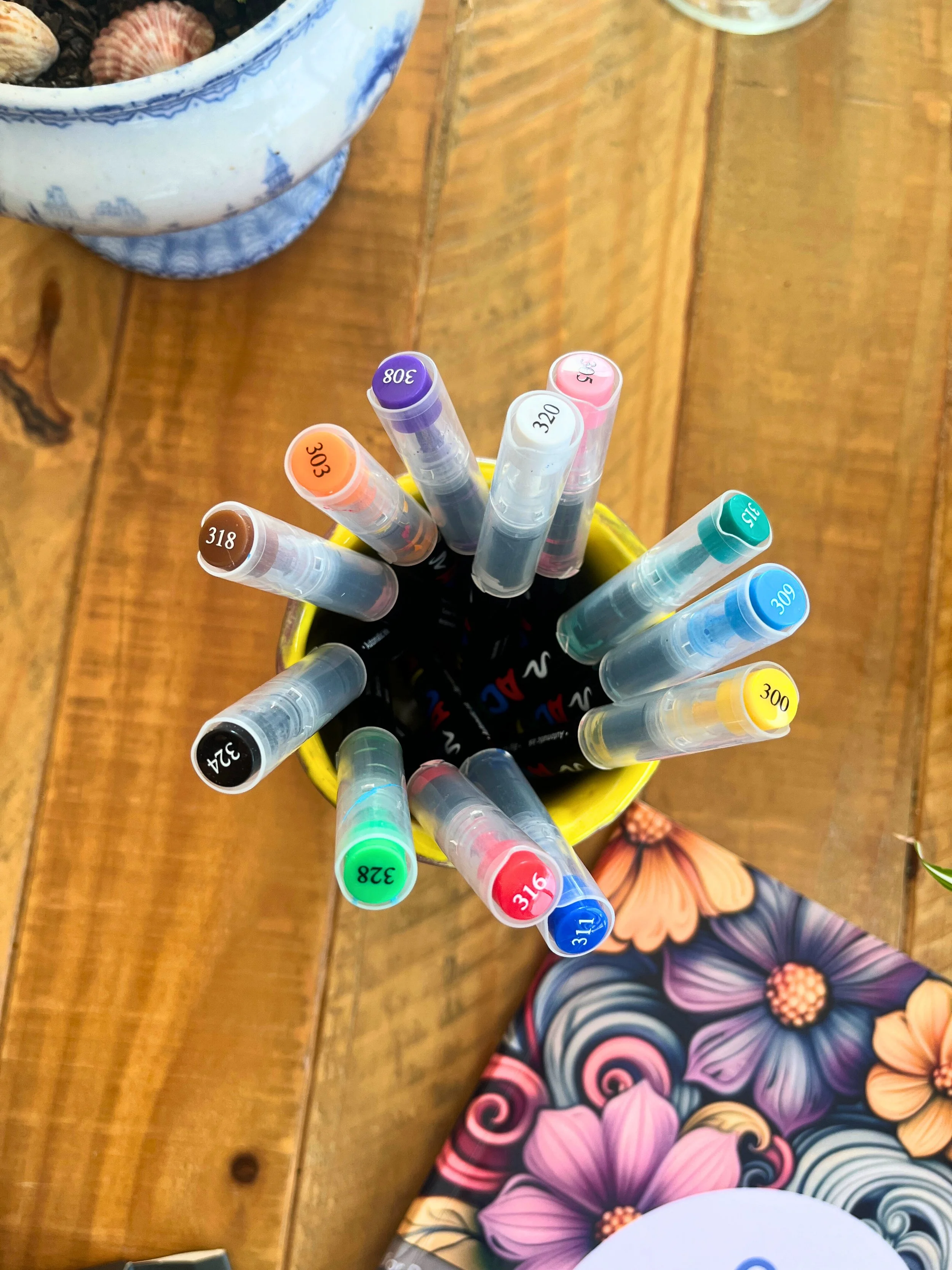 A top-down view of colorful markers in a yellow cup on a wooden table with a floral-patterned notebook nearby.
