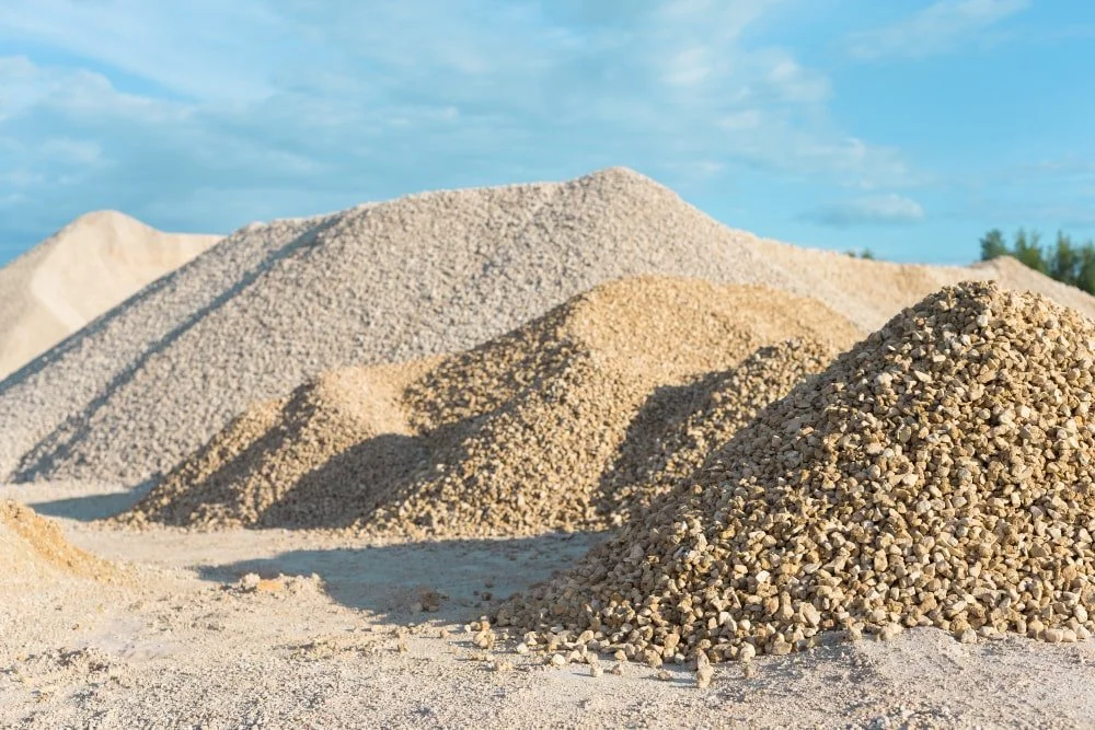 Multiple piles of gravel and sand outdoors on a sunny day with a blue sky and some clouds.