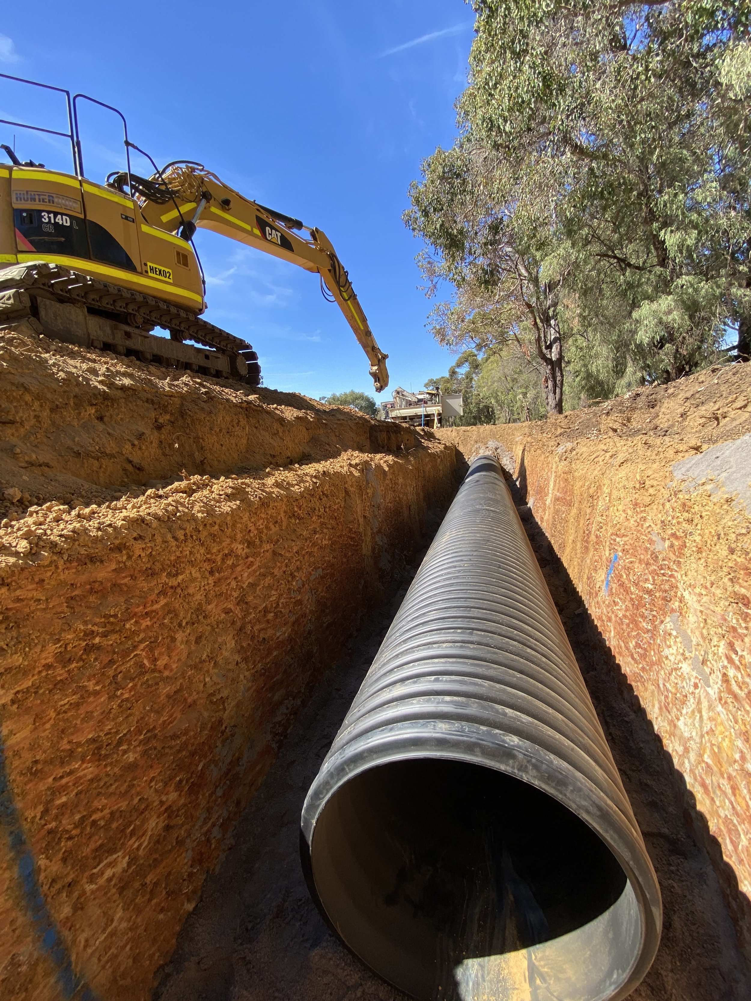 A construction site showing a large black pipe lying in a trench with an excavator above it, under a clear blue sky, and trees in the background.