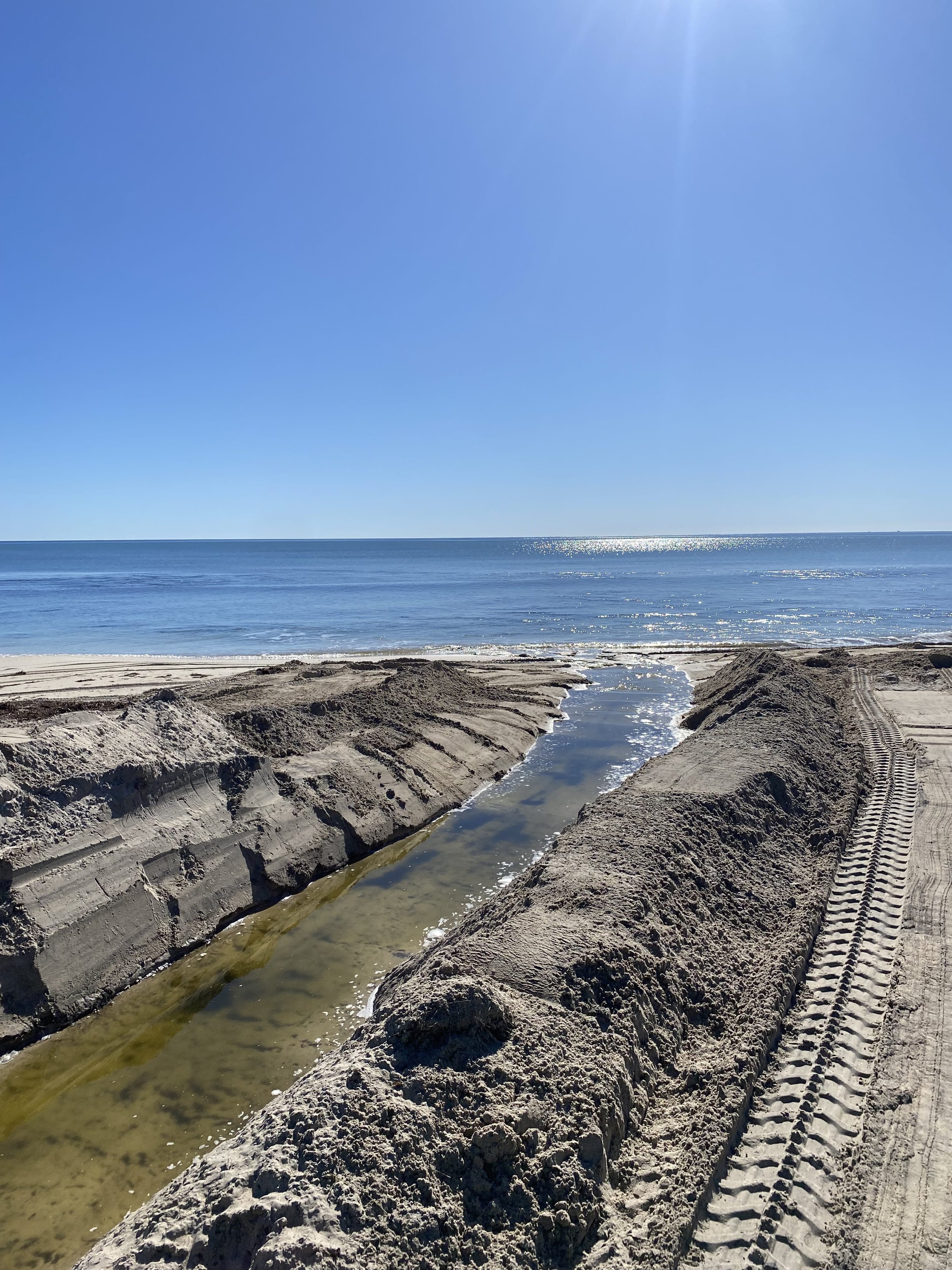 Construction on a sandy beach with a narrow water channel between sand mounds, pointing toward the ocean and clear blue sky.