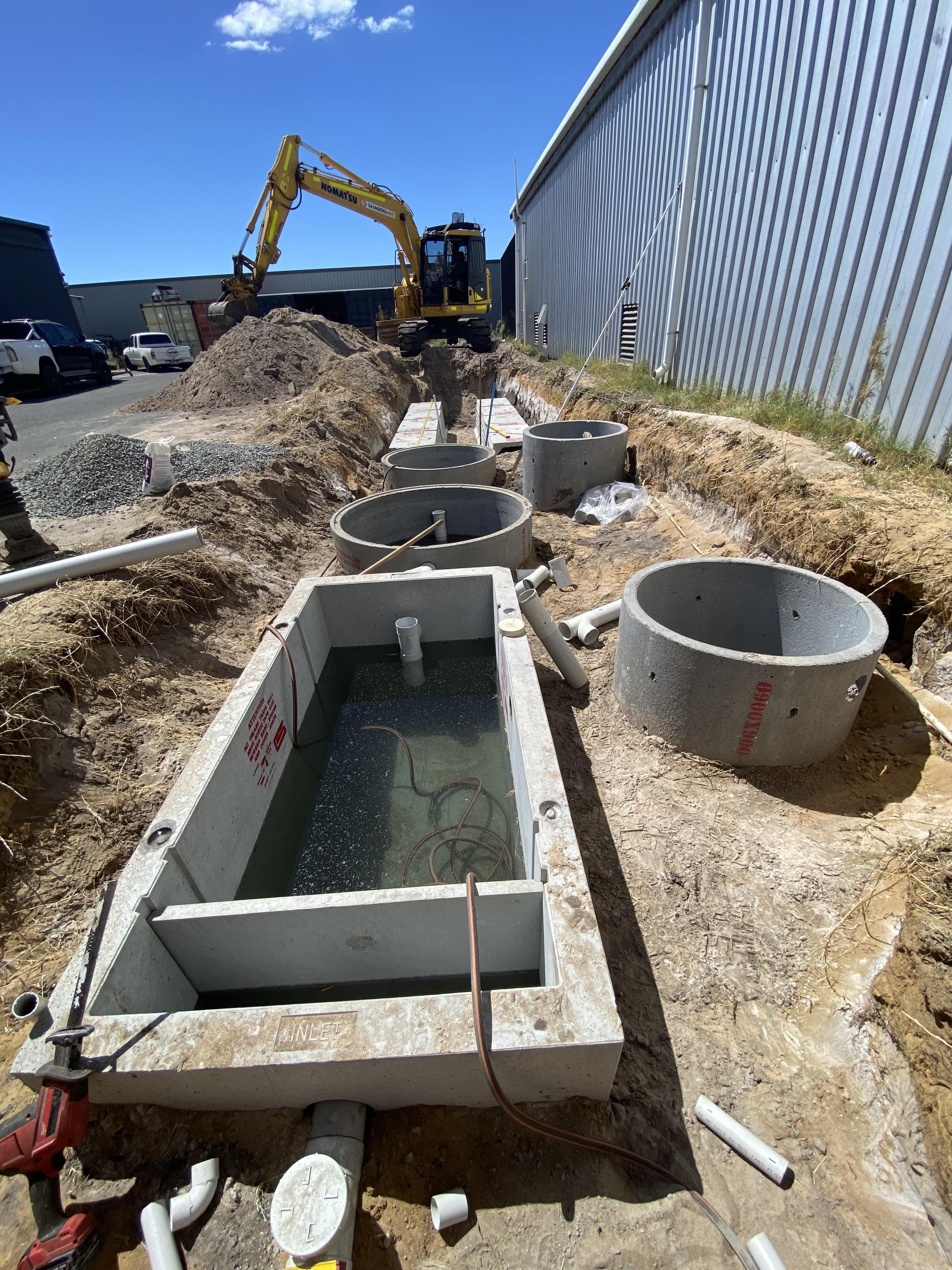 Underground utility construction site with concrete manholes, PVC pipes, and a yellow excavator working on the site under a blue sky.