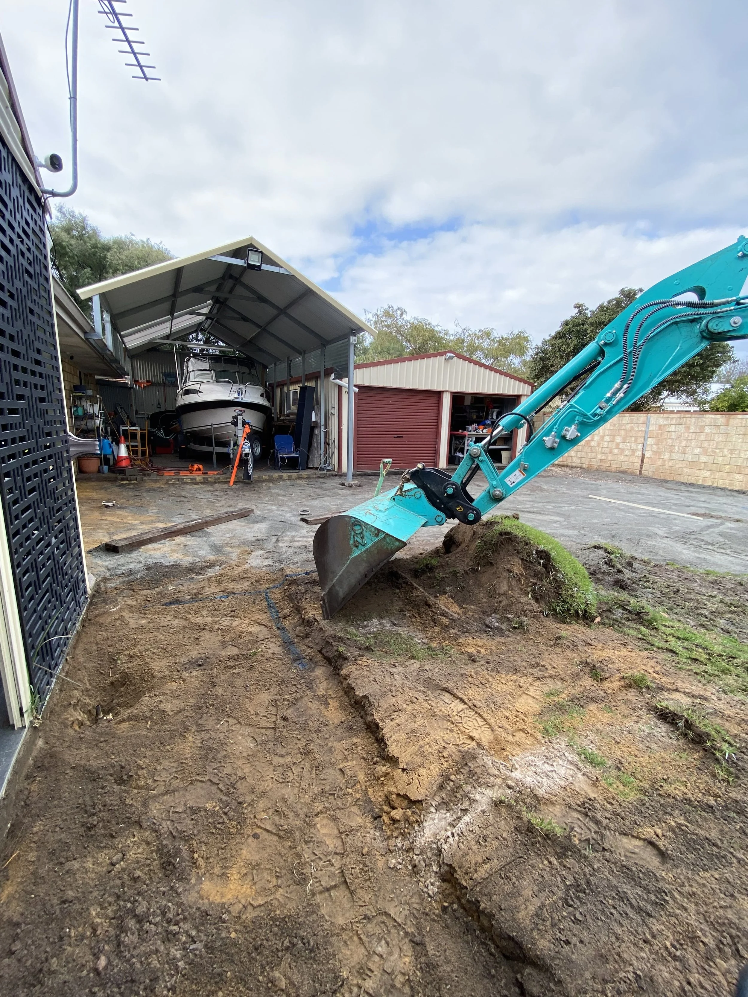 A small excavator digging out ground in a backyard with a boat stored in a metal shed in the background.