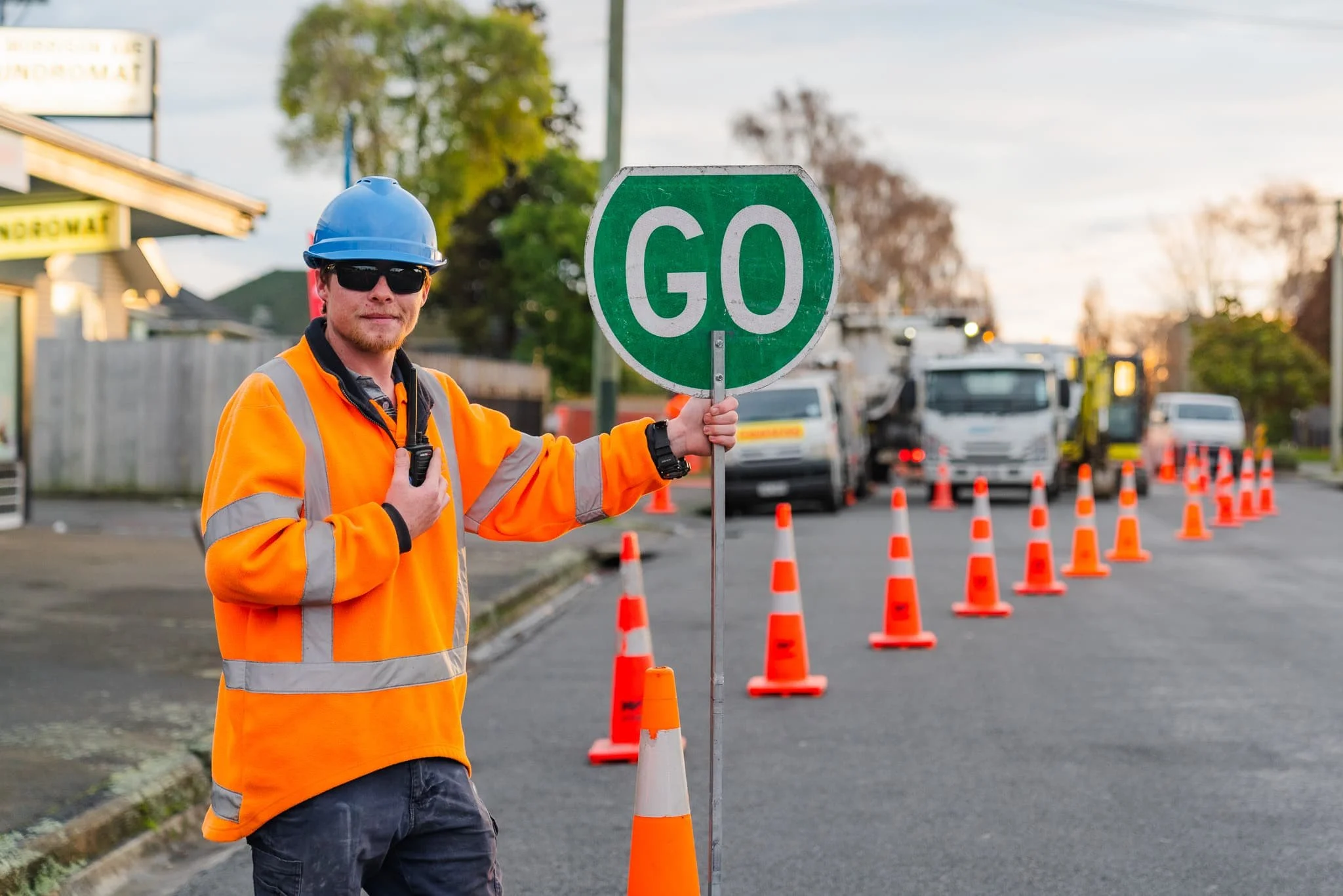 Construction worker wearing orange safety jacket, blue helmet, sunglasses, holding a STOP sign, standing on a street with orange traffic cones and trucks in the background.
