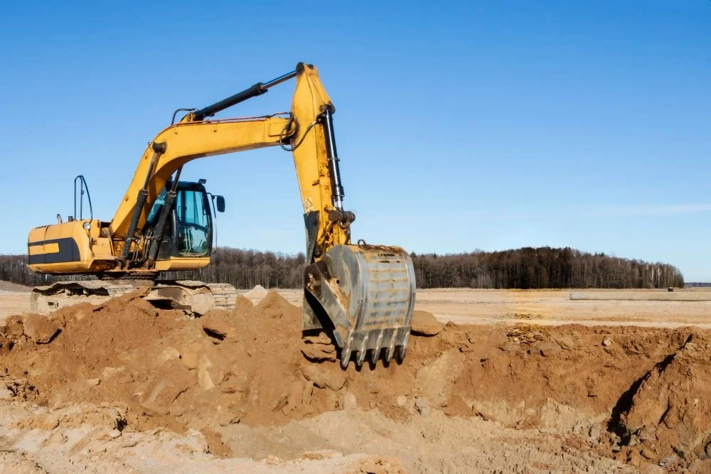 A yellow excavator digging in a dirt construction site with a clear blue sky and trees in the background.