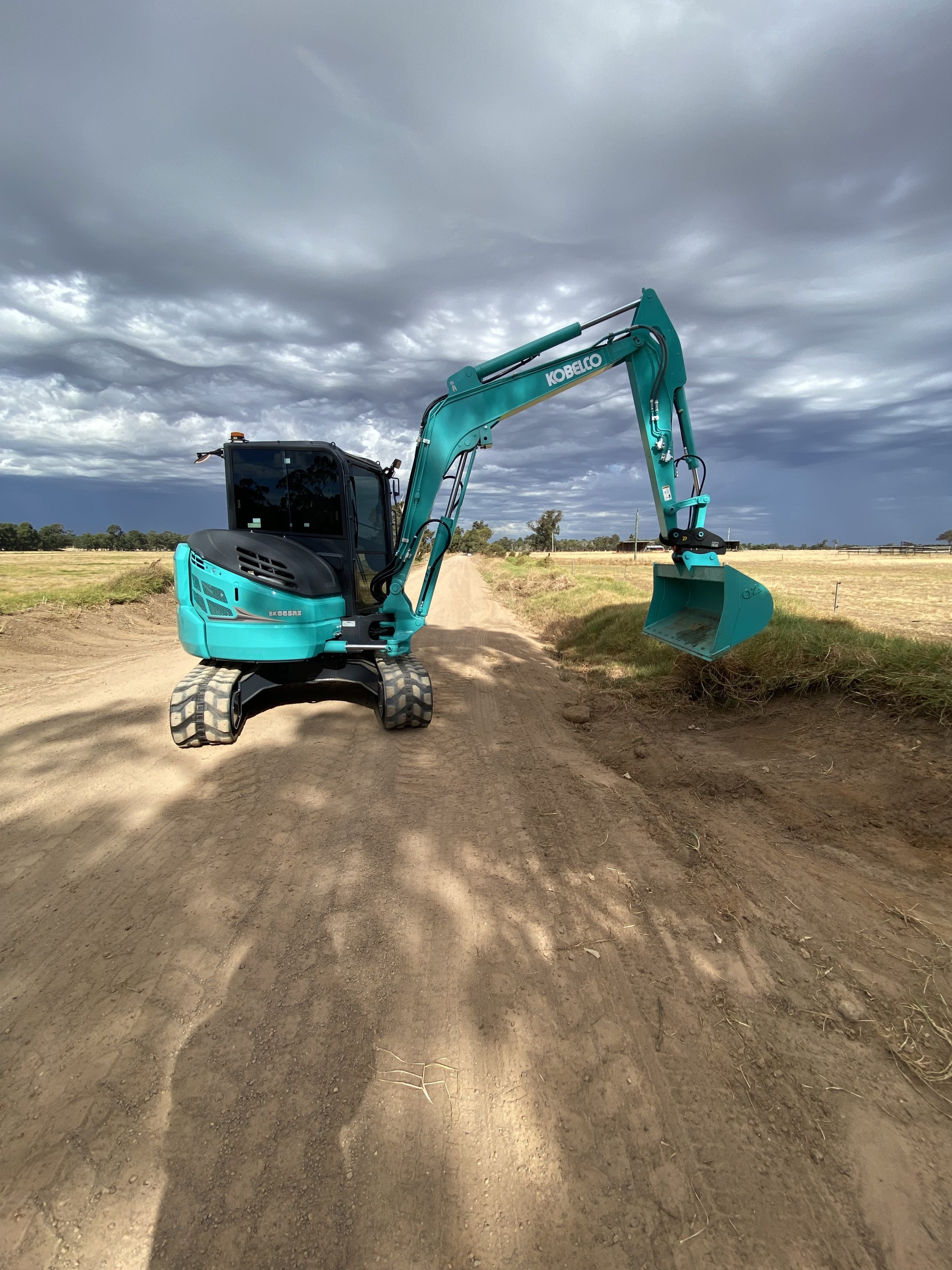A teal-colored mini excavator on a dirt road in a rural area under a cloudy sky.