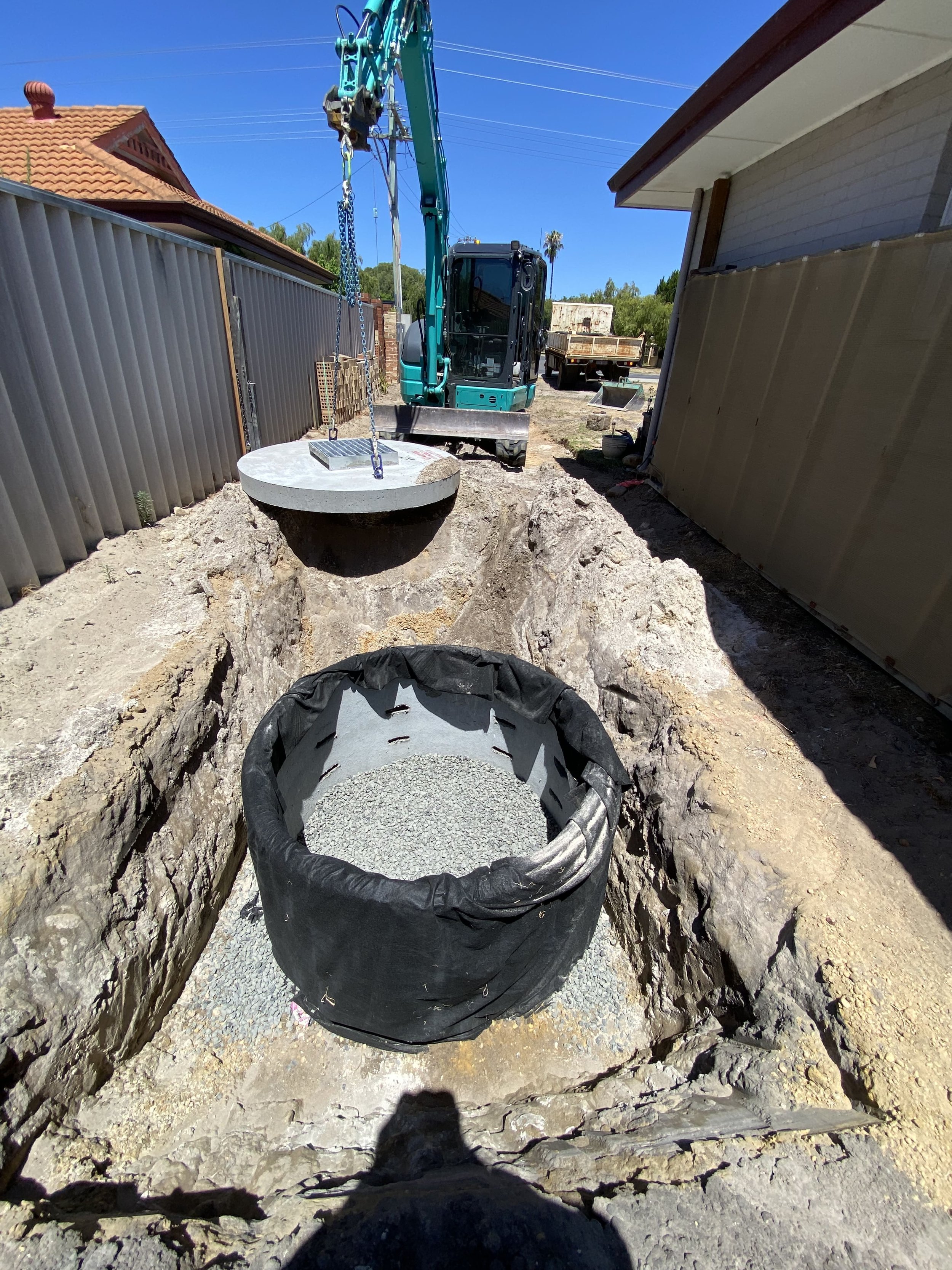 Construction site showing a small excavation with concrete work, a black container with gravel, and a teal excavator with a crane attachment lifting a concrete manhole cover, set in a sunny backyard with fences and neighboring houses.