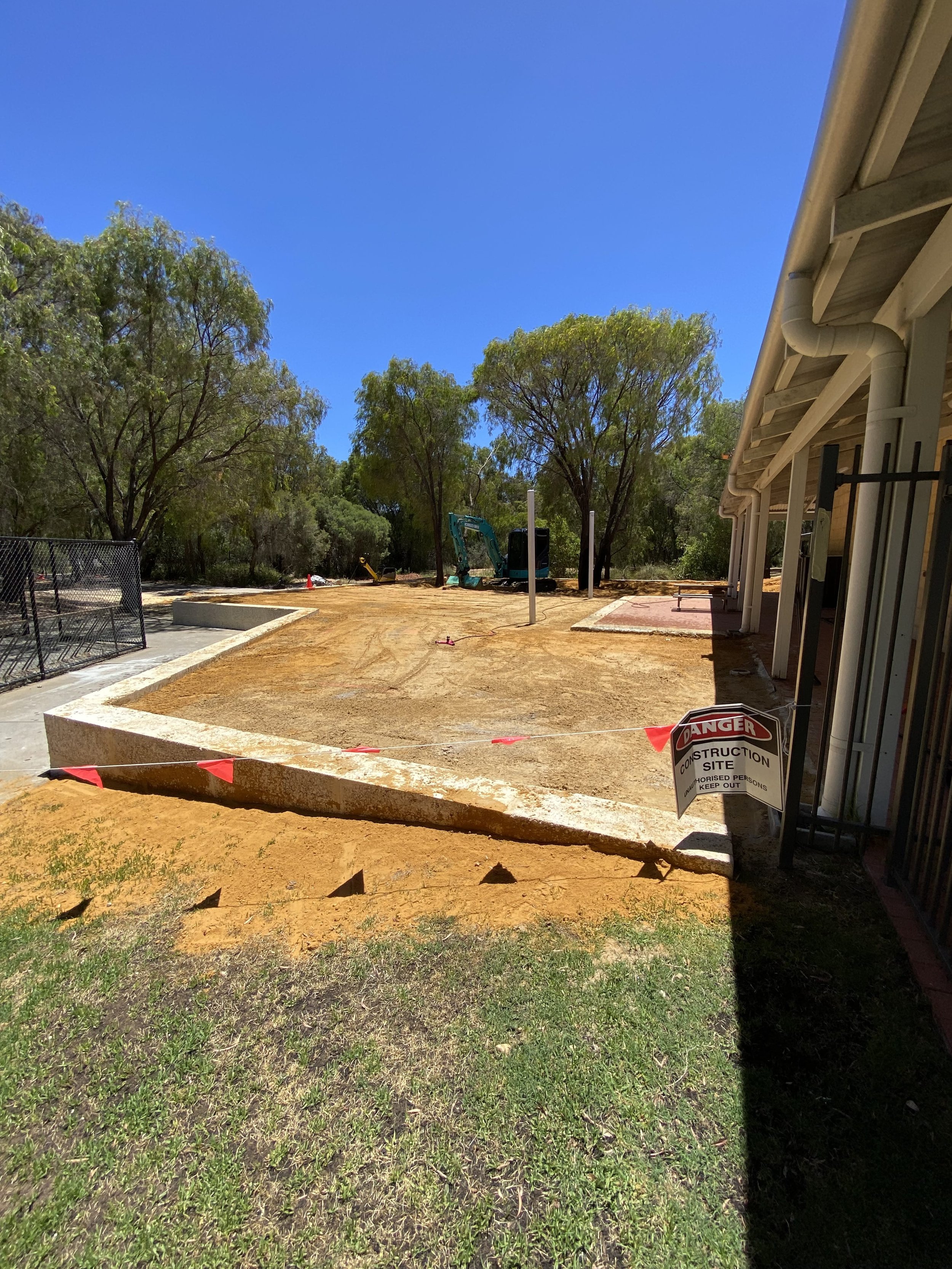 Construction site in a backyard with a partially built concrete foundation, enclosed by safety tape and a 'Danger Construction Site' sign, with trees and a house in the background under a clear blue sky.