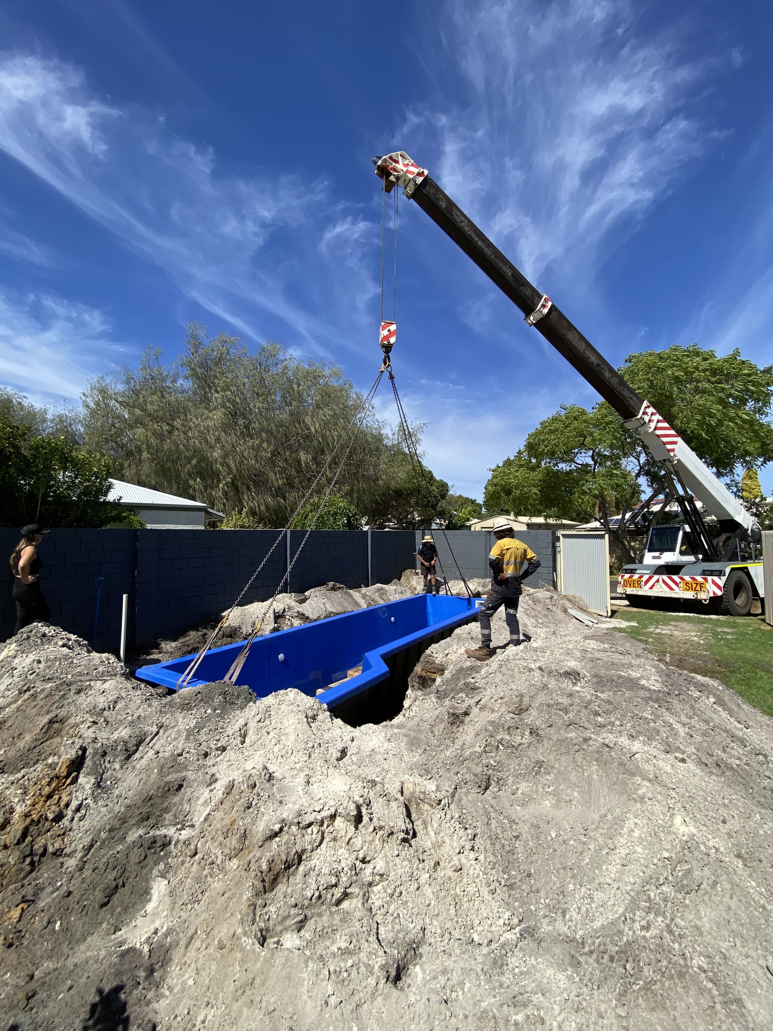 Construction workers install a blue swimming pool in a backyard, using a crane to lower it into a dug-out foundation amid dirt and rocks, with a blue sky overhead.