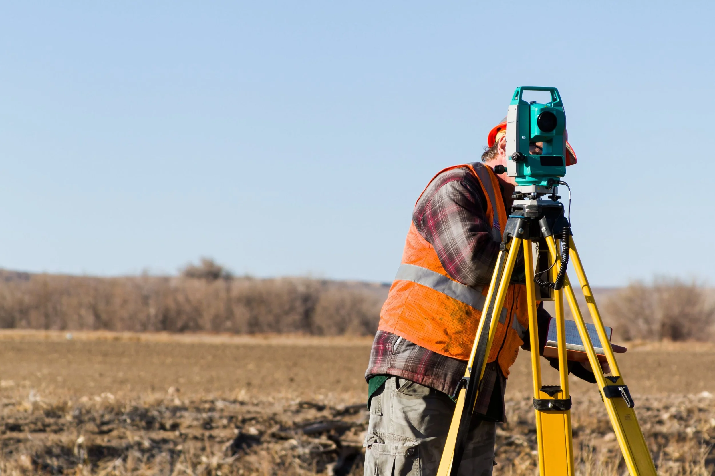 A person in a safety vest and plaid shirt using a surveying instrument on a tripod in an open field.