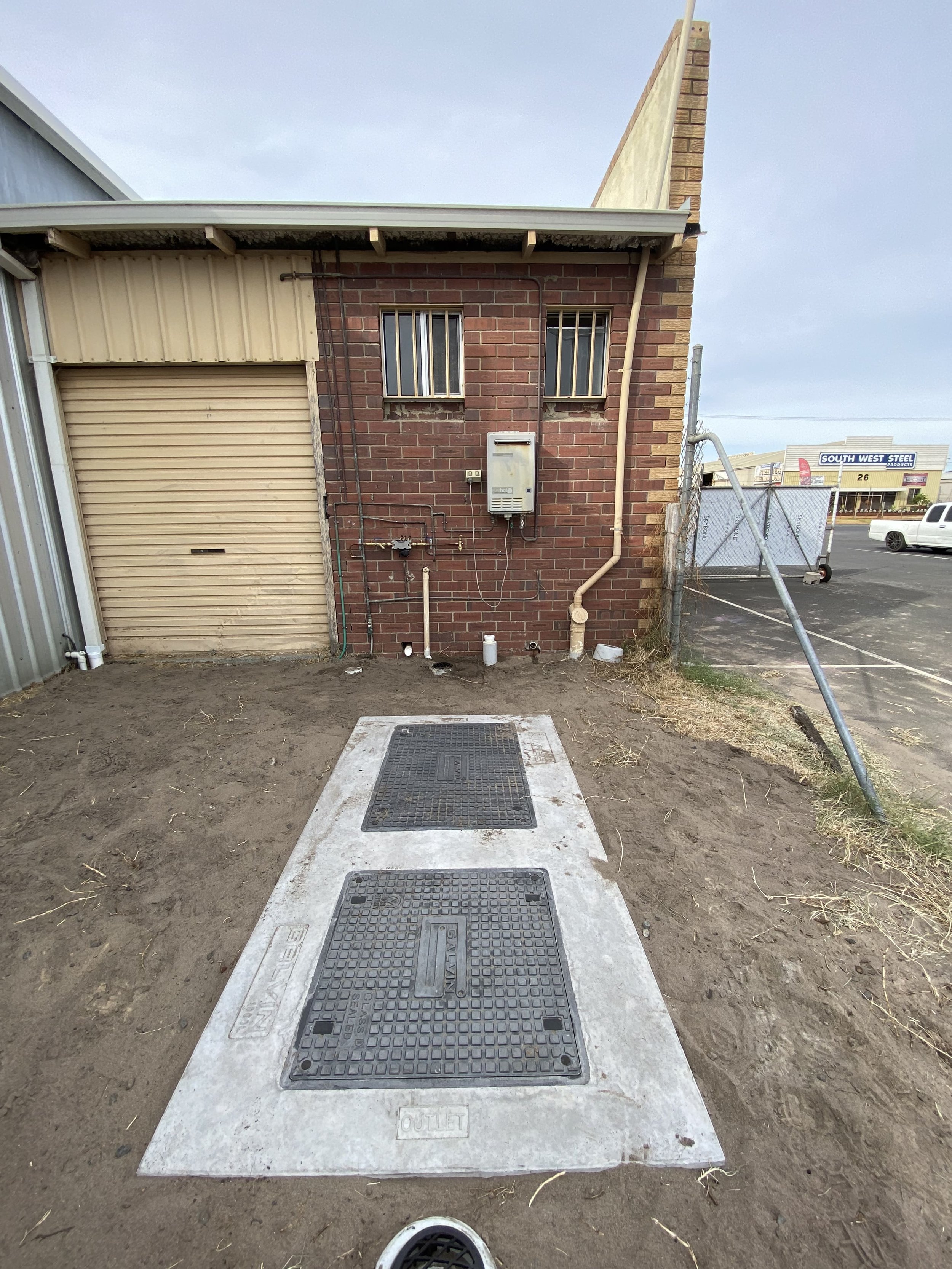 Concrete platform with two access hatches in front of a brick building with small barred windows and utility meters, surrounded by dirt and construction area.