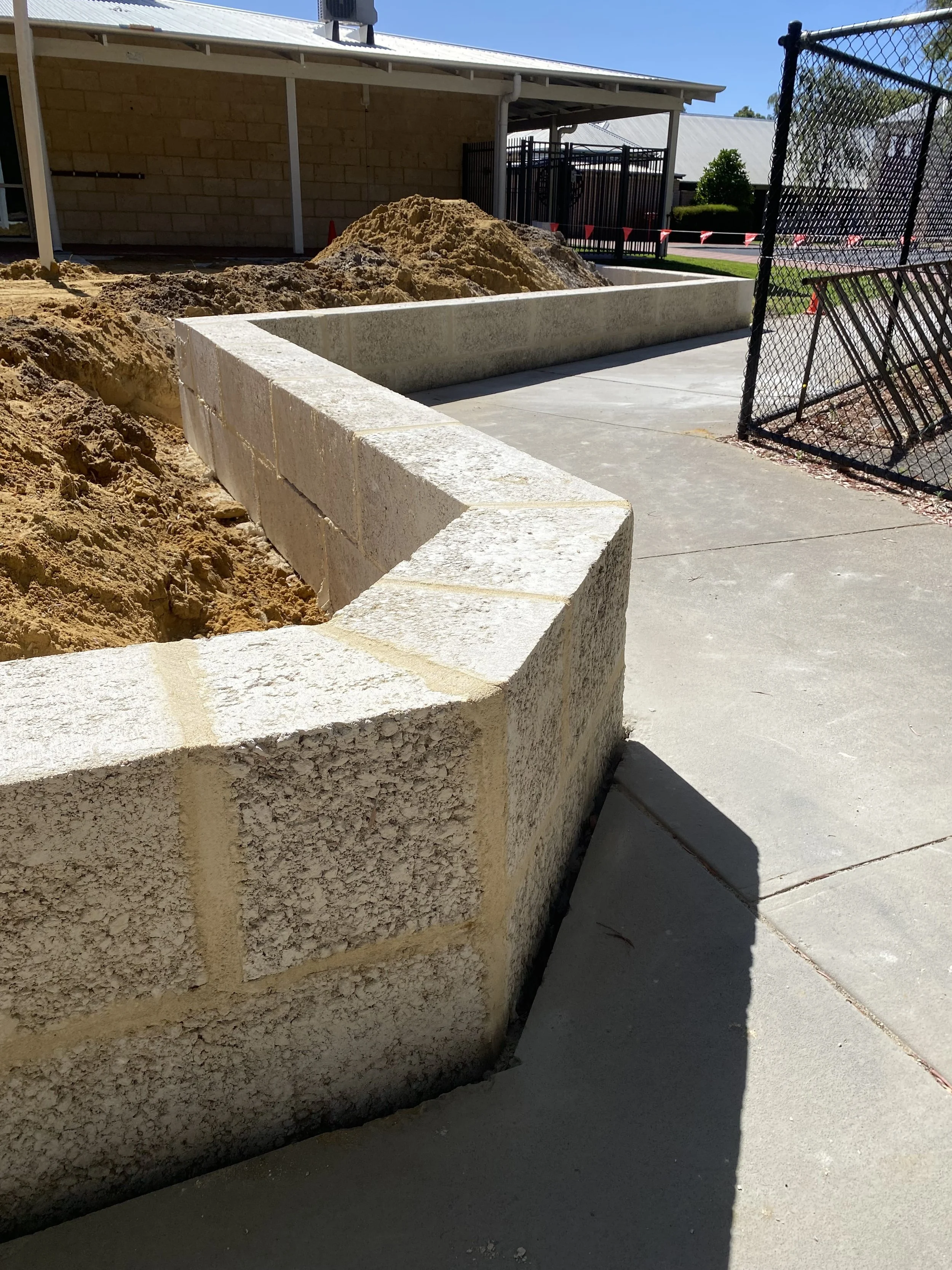 A concrete sidewalk with a stone retaining wall, construction site, dirt piles, and a chain-link fence in the background.