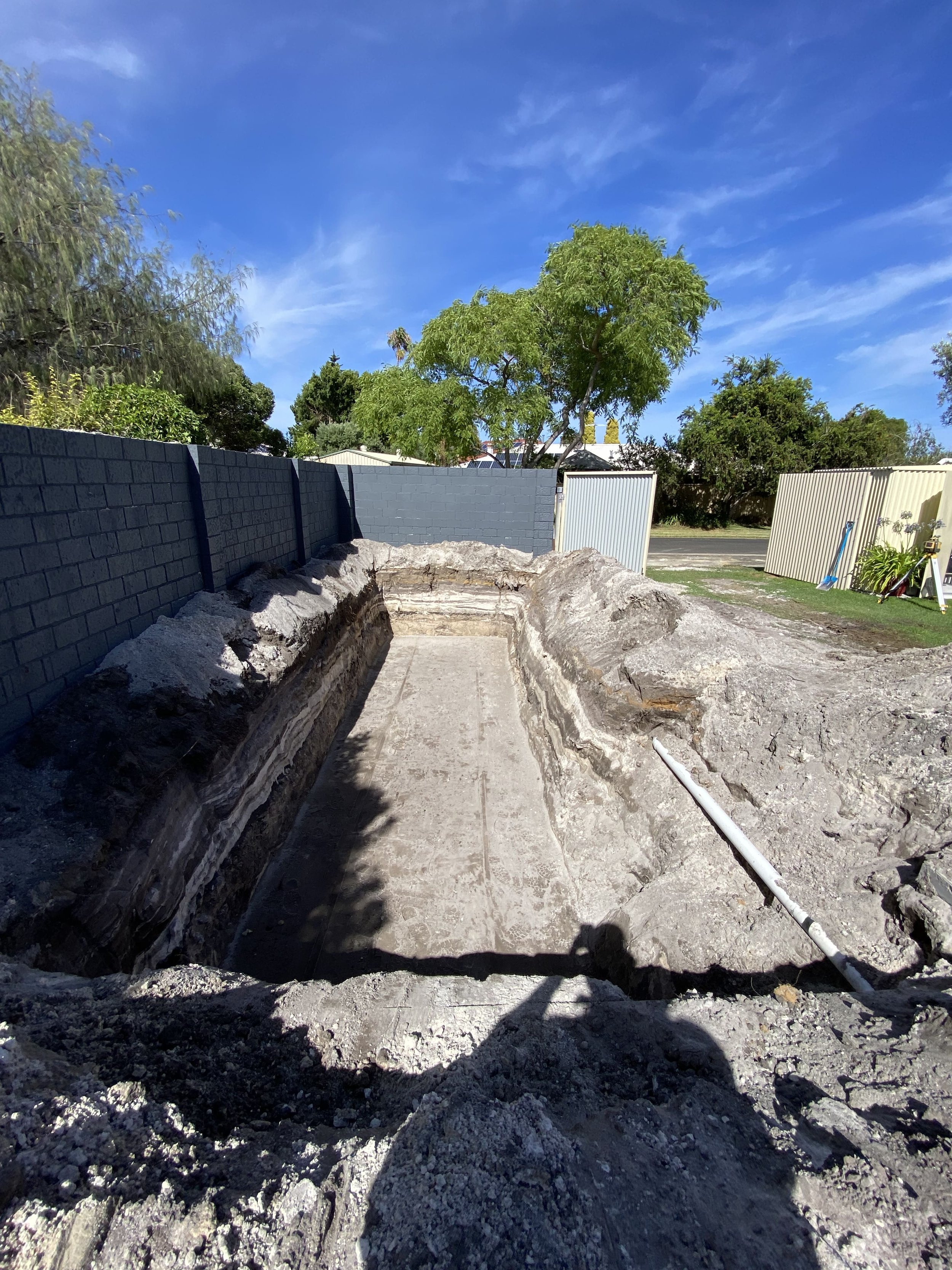 A construction site with a dug trench, a gray brick wall on the left, and a bright blue sky with some clouds. There are trees and a fence in the background.