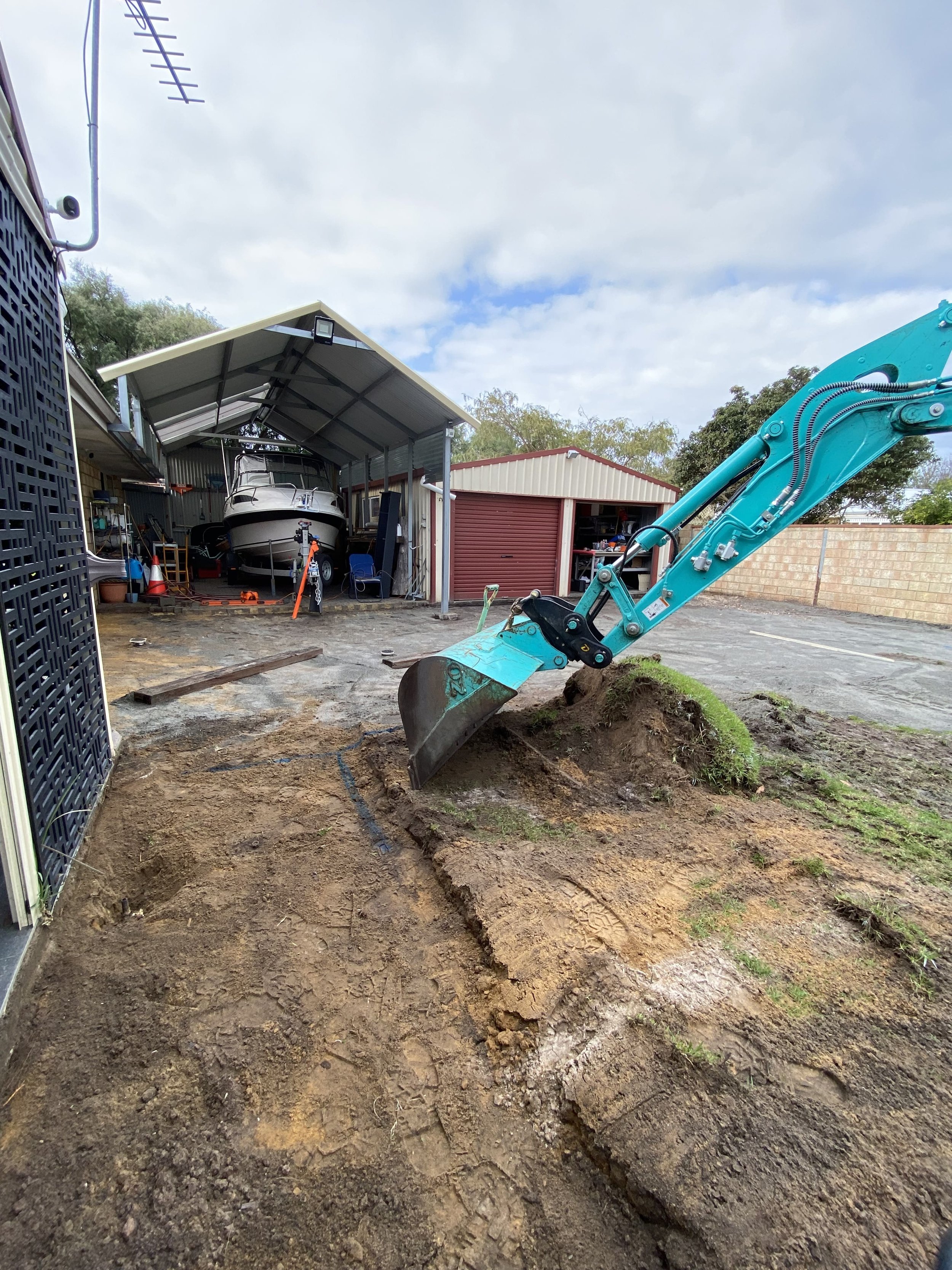 A small excavator digging a trench in a backyard near a storage shed with boats, boats, and garage in the background.