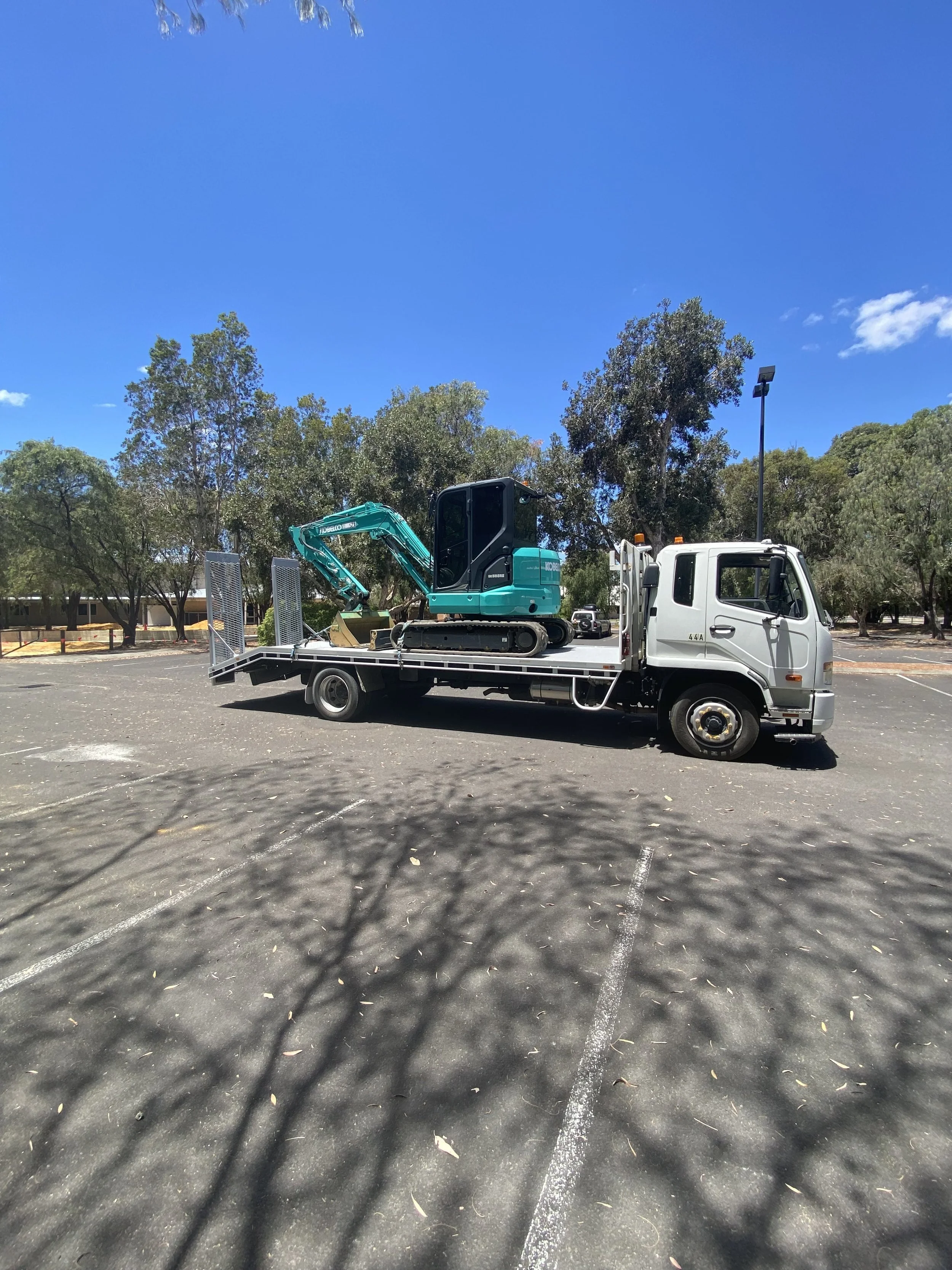 A white flatbed tow truck parked in an empty parking lot with a small teal excavator loaded on it. Trees are in the background under a clear blue sky.