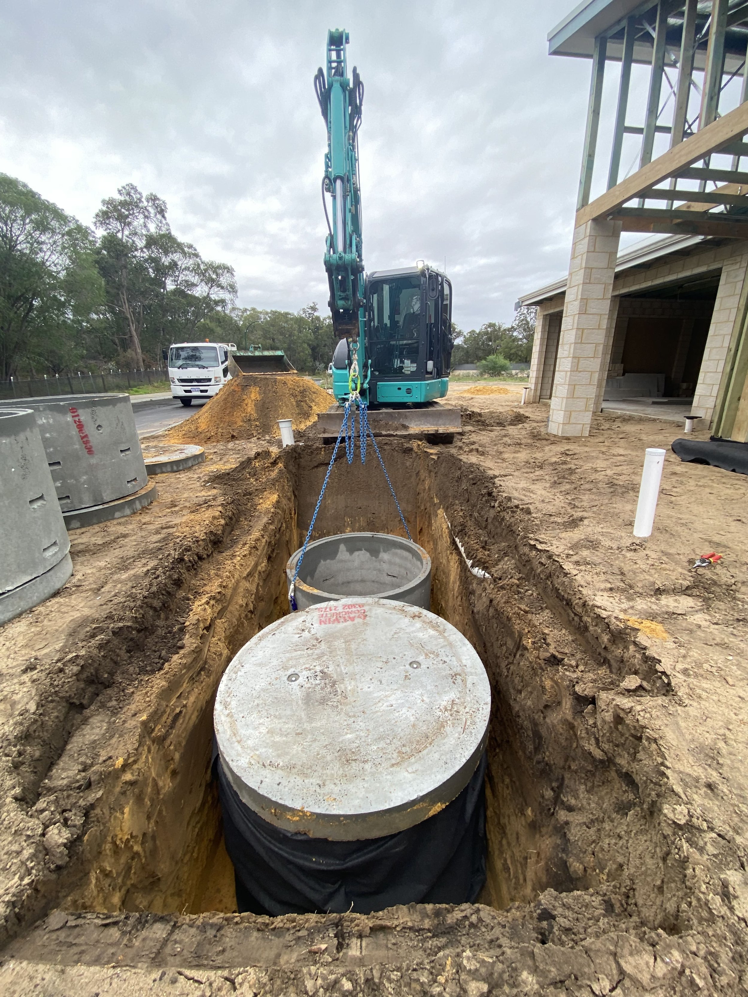 Construction site with a small excavator installing underground utility pipes, one pipe is lifted by a crane hook.