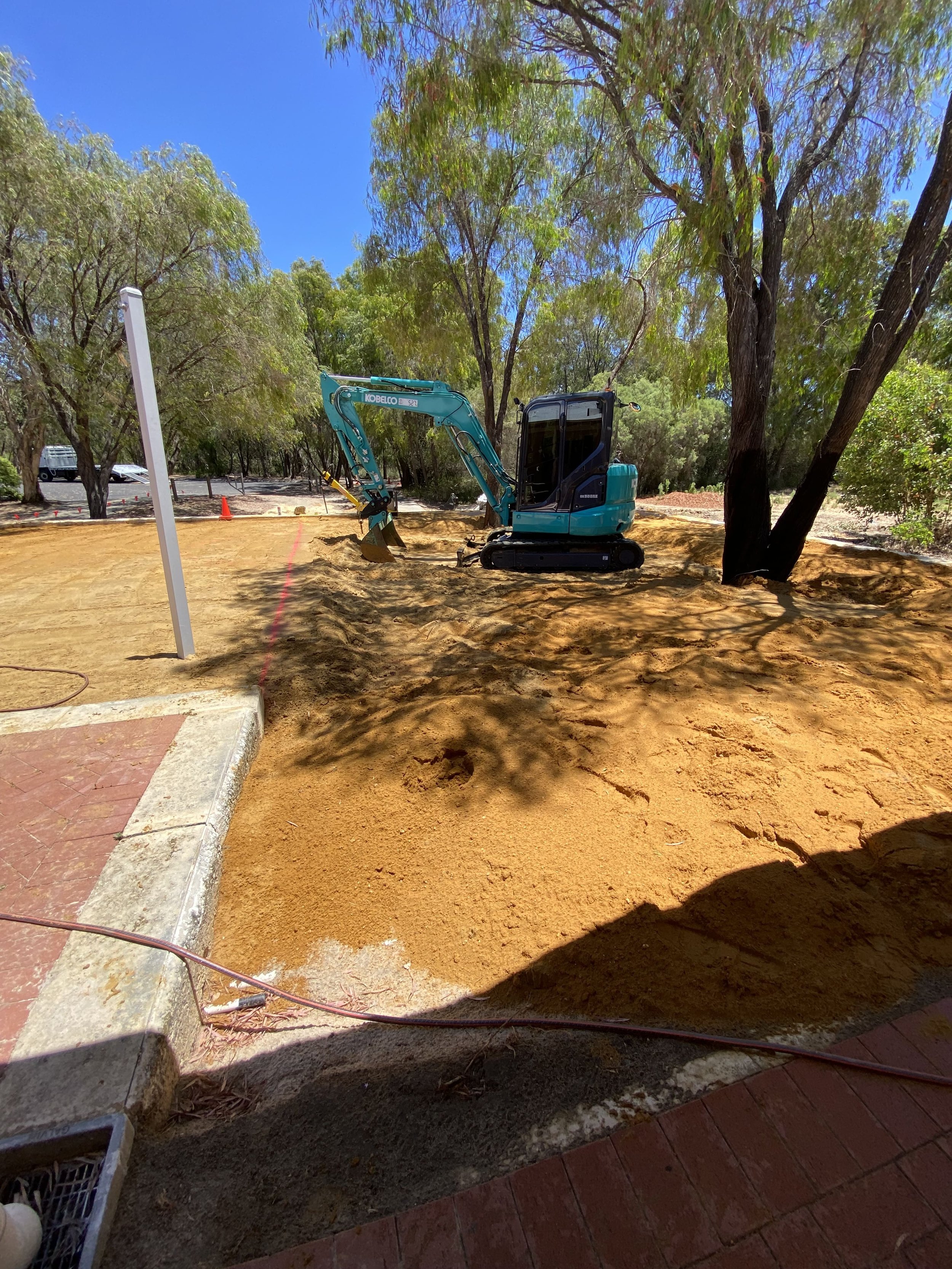 Construction site with a small blue excavator digging on a dirt area surrounded by trees under a blue sky.