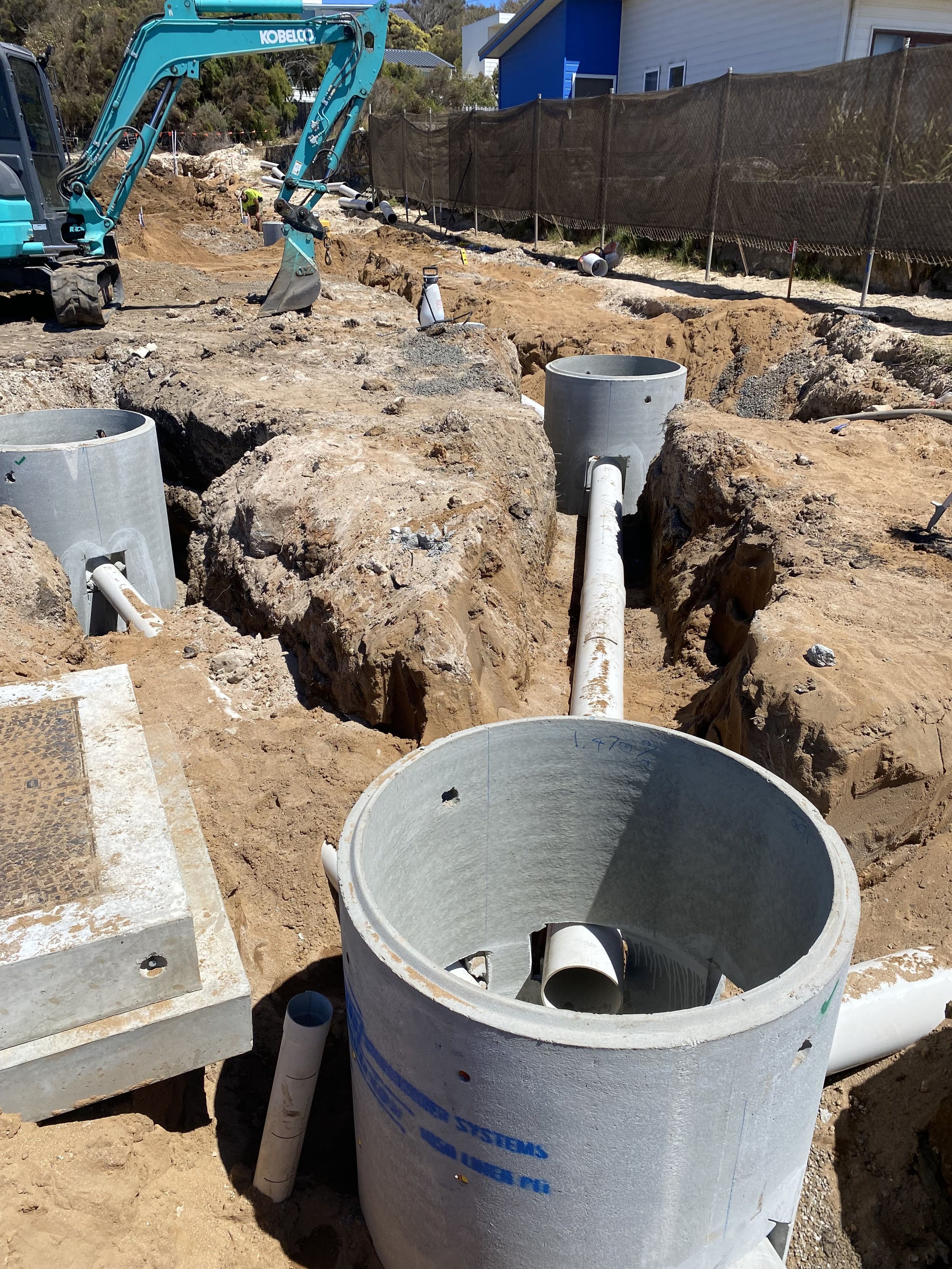 Construction site with underground utility pipes, large concrete pipes, and excavation equipment under a clear sky.