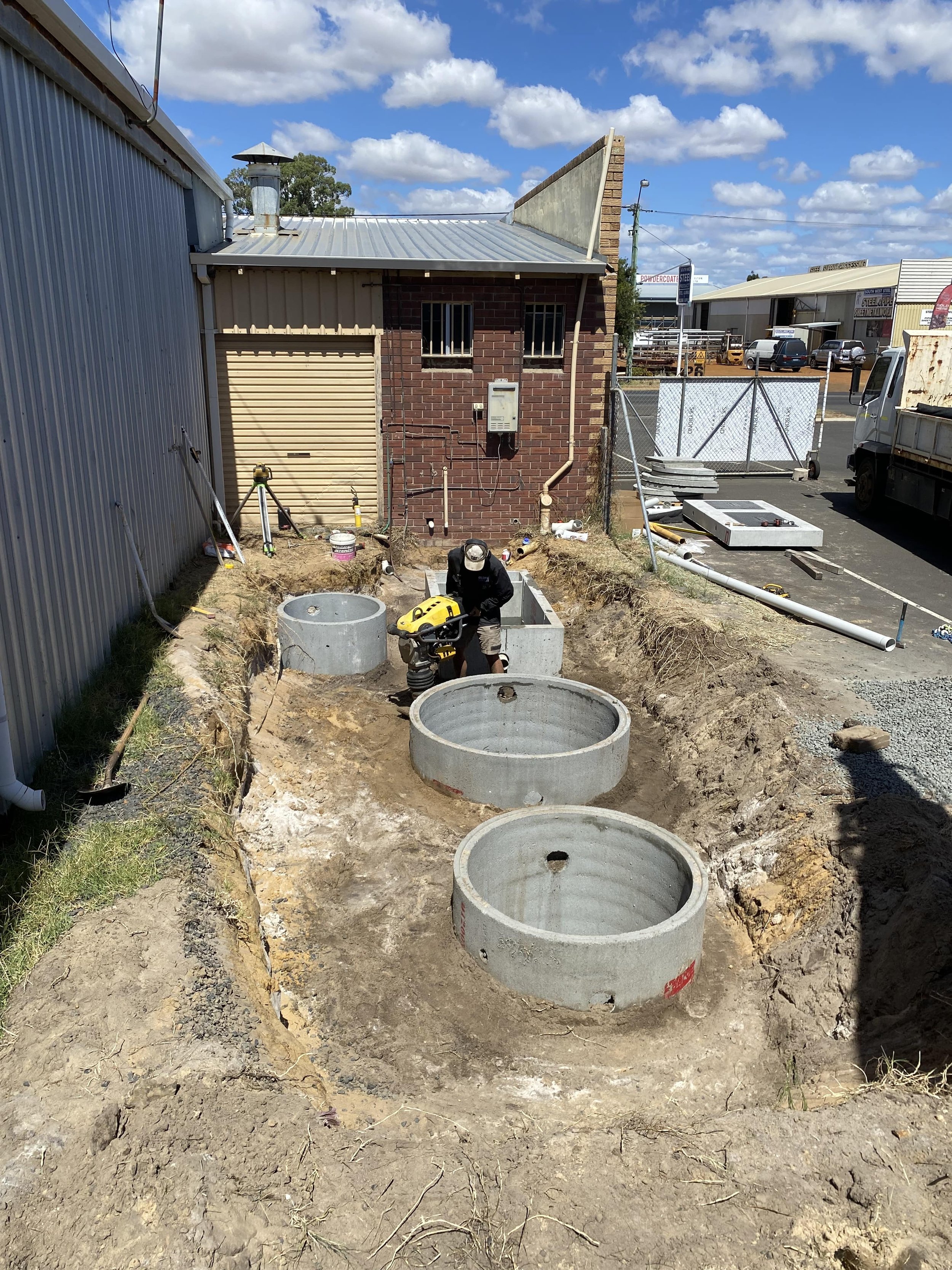 Construction worker installing underground concrete sewer or drain manholes at a building site. The site is dug out with several large concrete cylindrical structures placed in the ground.