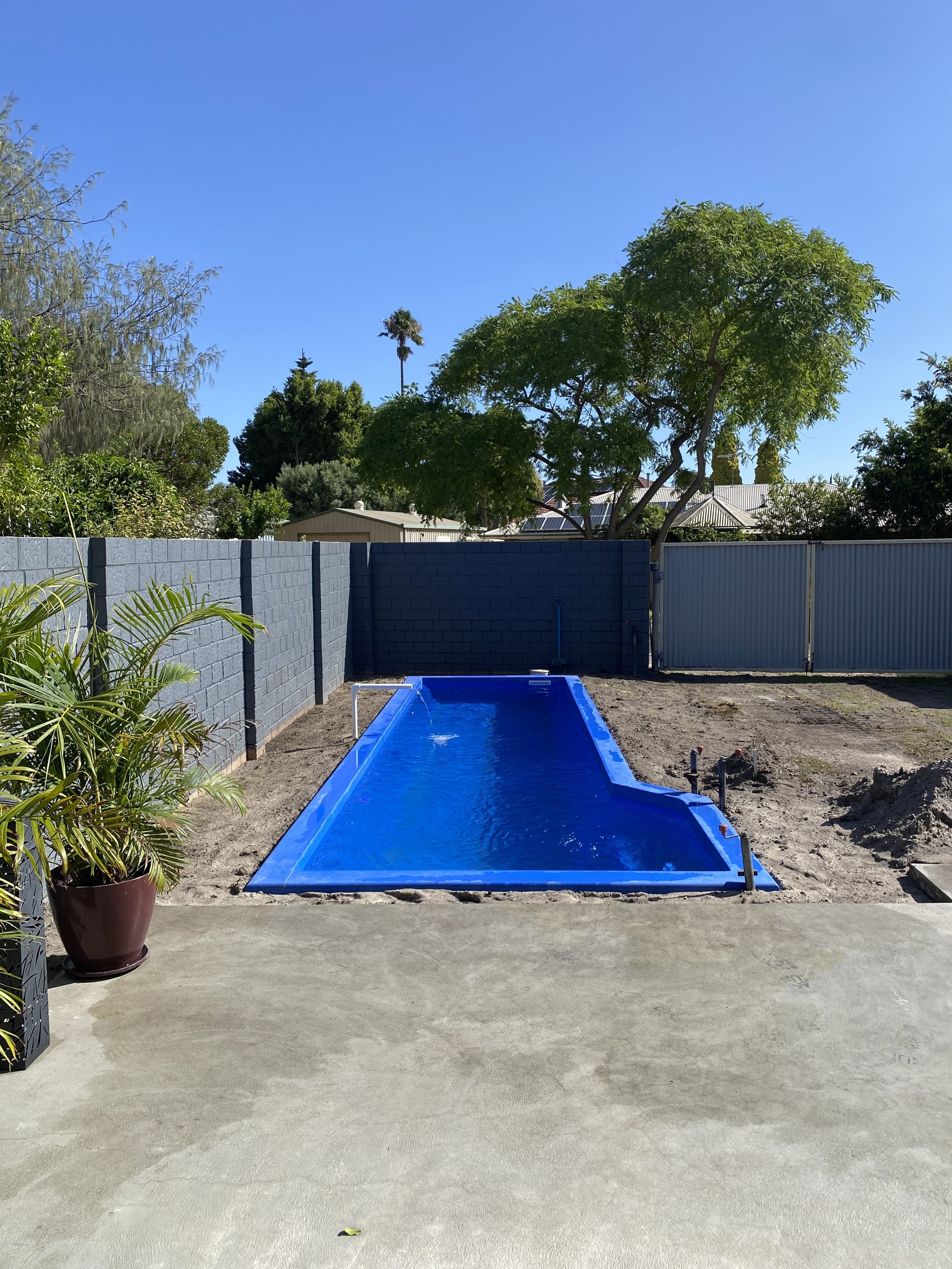 A backyard with a new, empty in-ground swimming pool with blue liner, surrounded by dirt and a concrete patio, enclosed by a gray fence, with trees and clear blue sky in the background.