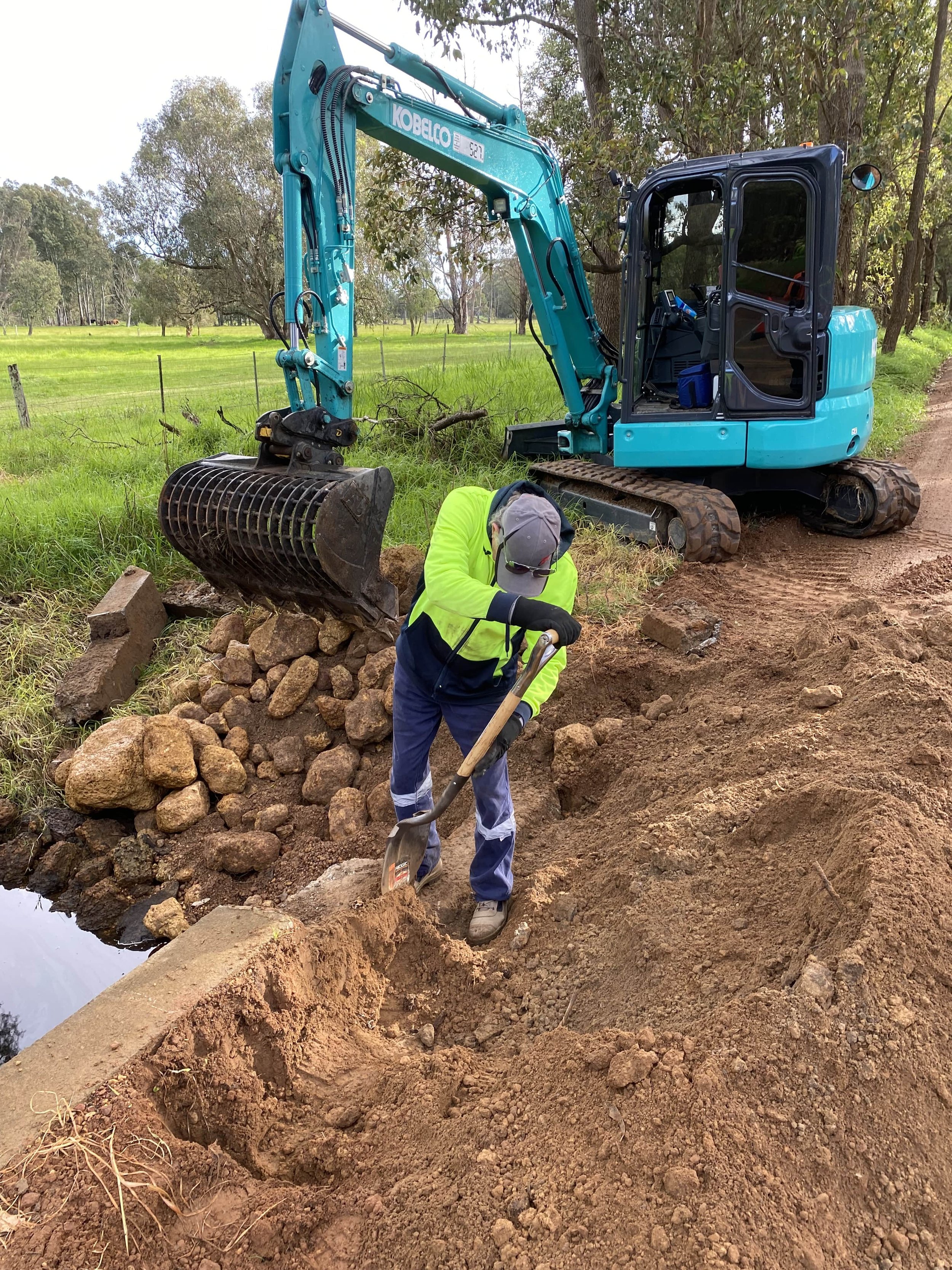 A person in a high-visibility jacket and cap using a shovel to dig a trench next to a waterway, with a blue excavator parked on the dirt nearby.