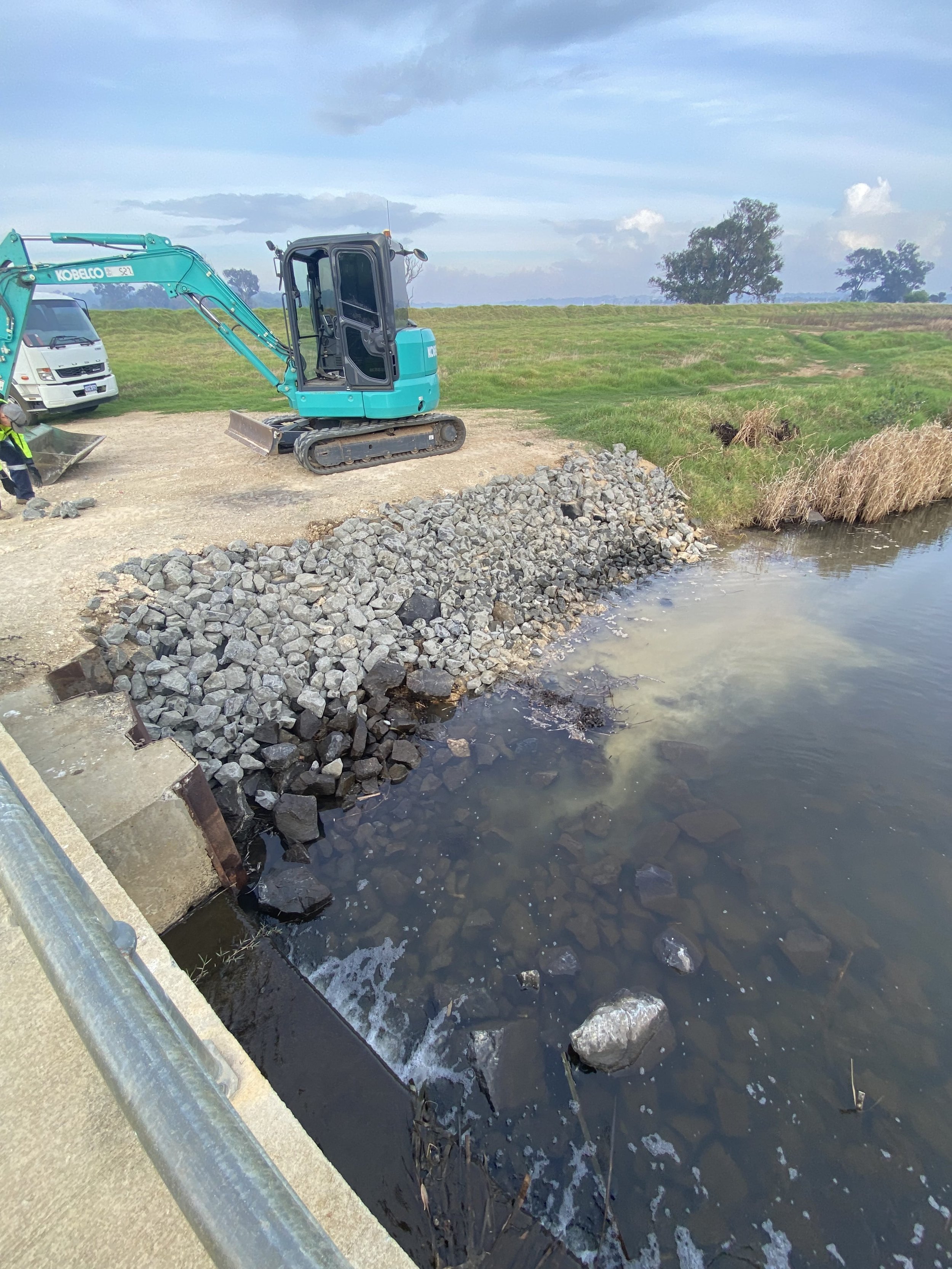 Construction equipment and rocks on a bank by a body of water, with a grassy field and trees in the background.