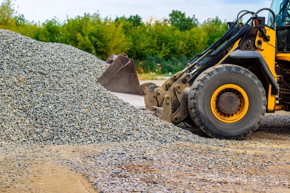 A yellow bulldozer with black hydraulic arms and large black tires moving a pile of gravel on a construction site with green trees in the background.