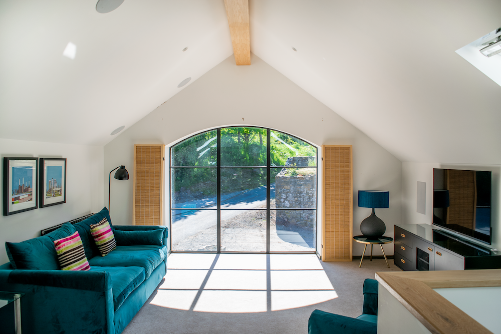 Living room with large arched window, teal sofa with colorful pillows, wall art, side table with blue lamp, and TV on a black stand.